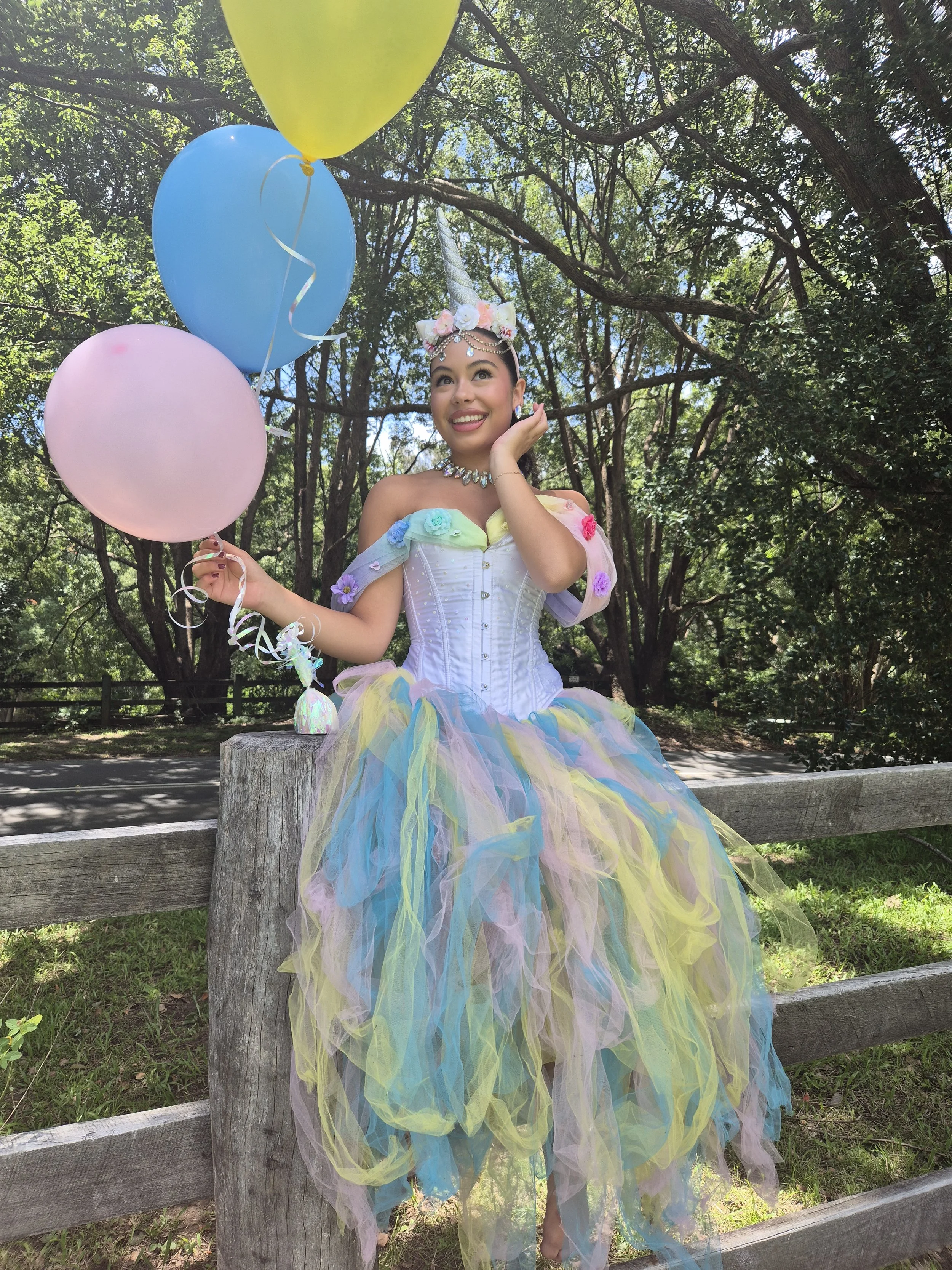 A young woman dressed as a unicorn fairy princess, wearing a glittering unicorn horn headpiece, a pastel-colored tulle tutu, and a colorful off-shoulder top with floral and gem accents, standing outdoors against a background of trees, holding pastel-colored balloons in pink, blue, and yellow.