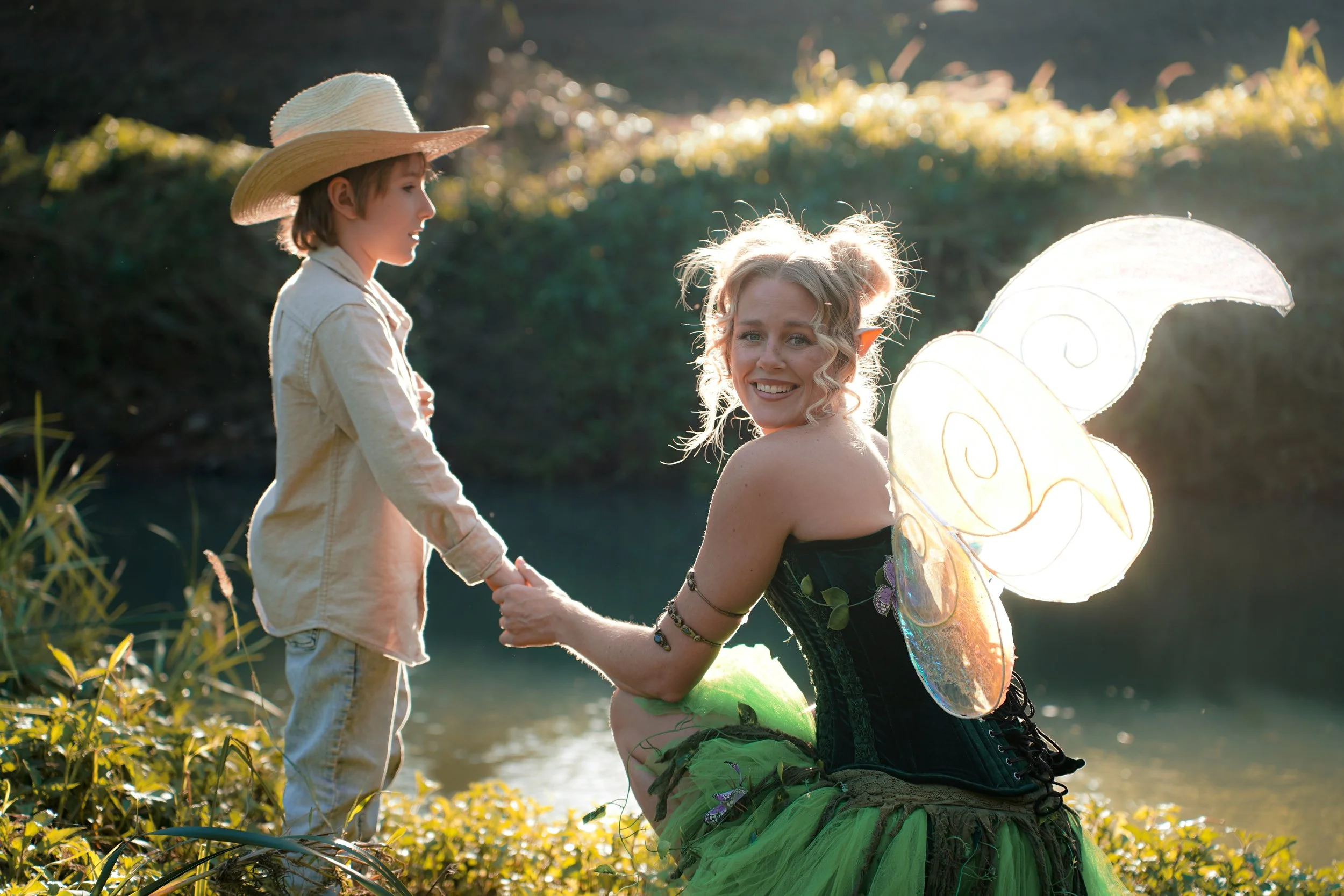 A woman dressed as a fairy with butterfly wings, a green skirt, and fairy ears is kneeling by a riverbank, holding hands with a young boy wearing a large sun hat and a beige jacket, as they enjoy a moment in a natural outdoor setting during sunset.