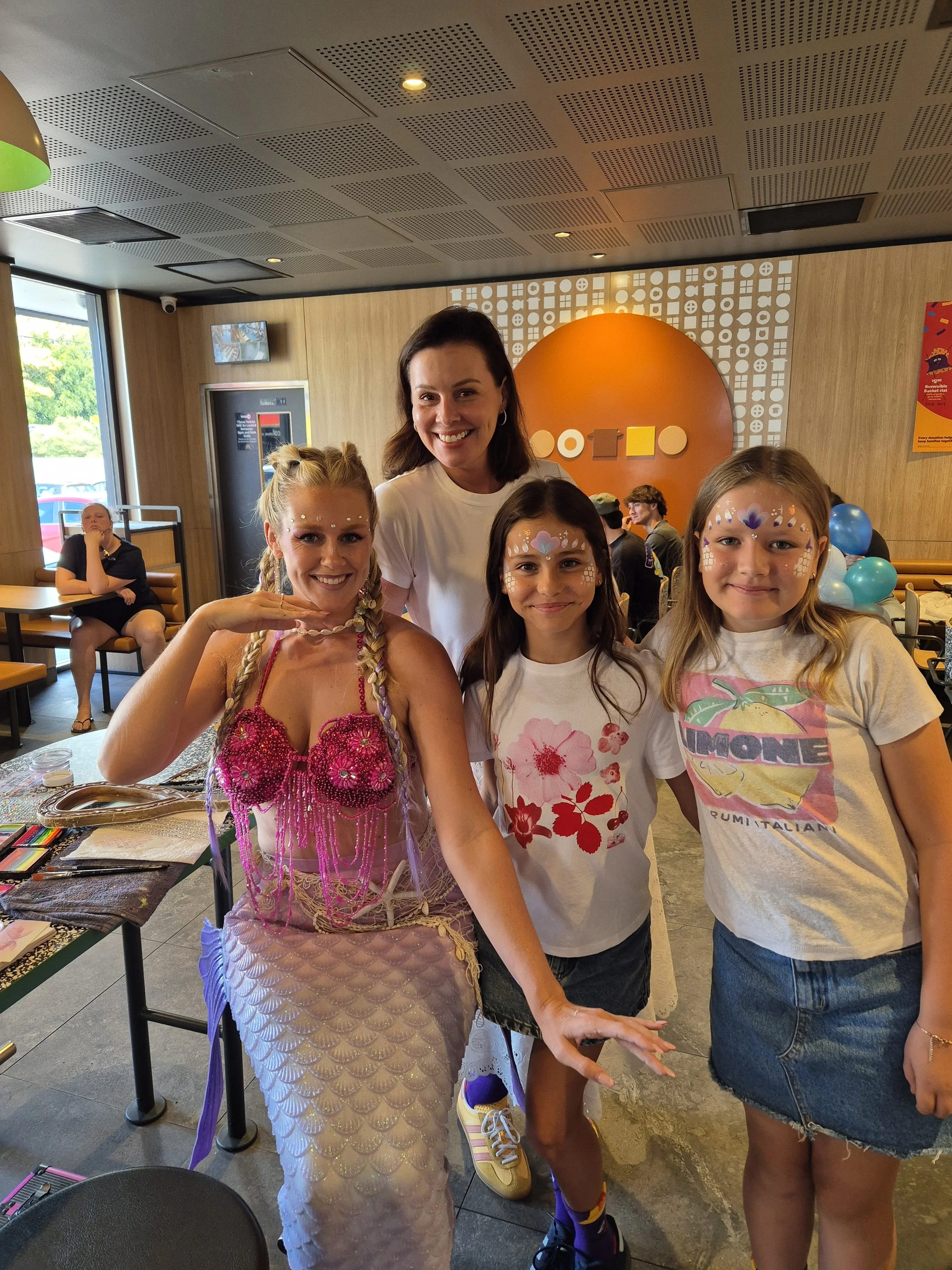 Four women are smiling inside a restaurant or cafe, with three of them wearing face paint with colorful patterns and accessories, while one woman in the background is seated at a table and looking at her phone.
