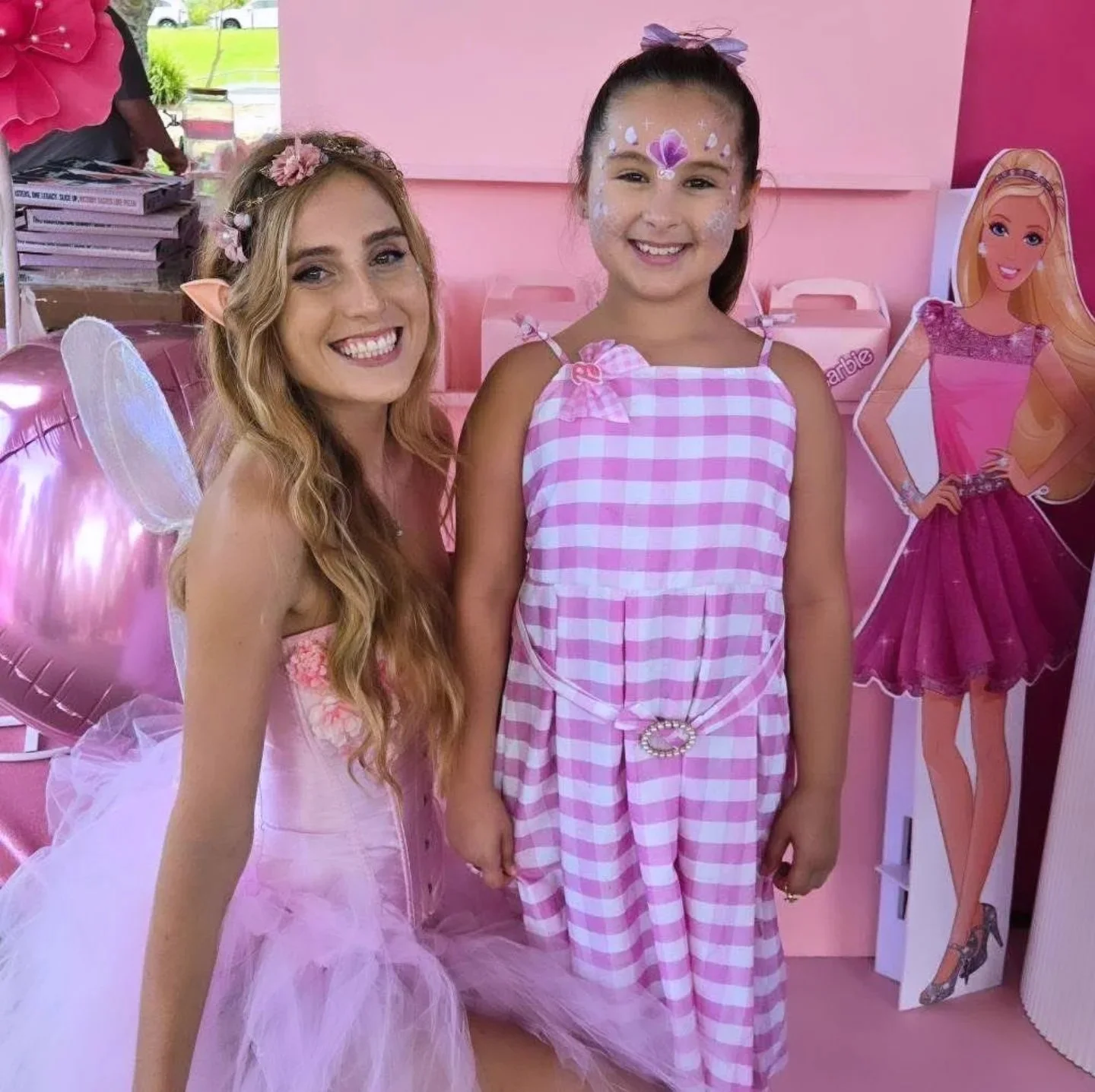 A woman dressed as a fairy with fairy wings and a pink tutu is smiling next to a girl in a pink and white checkered dress with face paint, standing at a pink-themed birthday party with a Barbie cardboard cutout in the background.