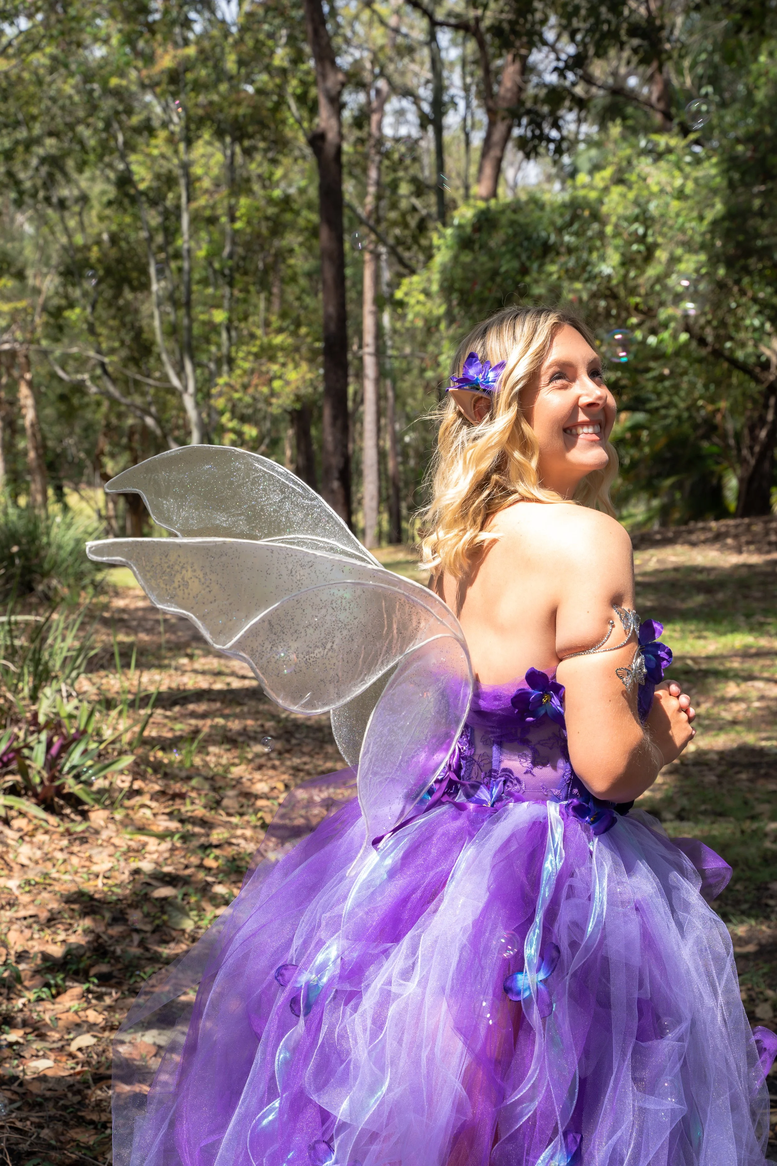 A woman dressed as a fairy with purple and lavender tulle dress, shimmering fairy wings, and purple flowers in her blonde hair, standing outdoors in a wooded area, smiling and looking to the side.