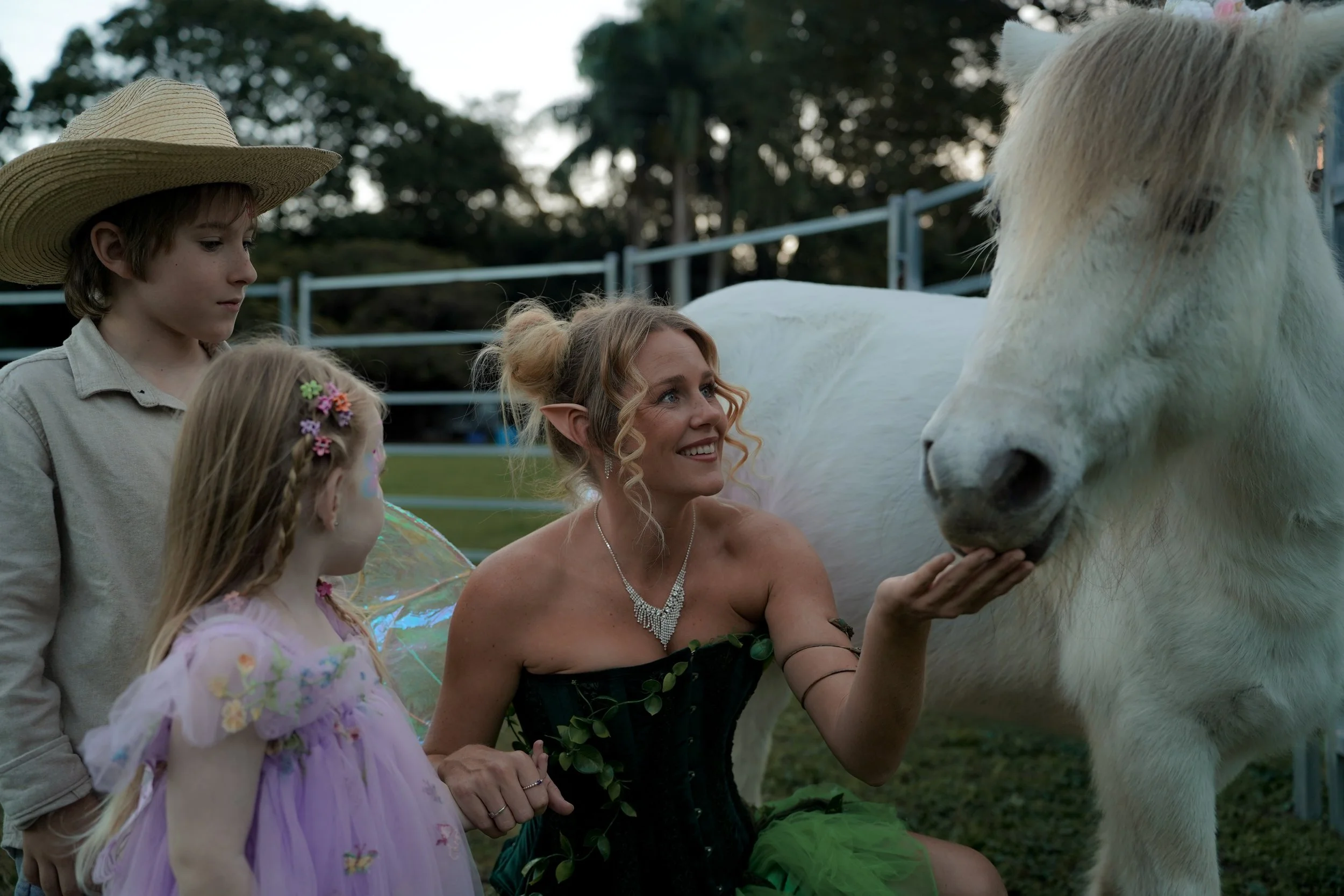 Young girl in a purple dress with flower accents and fairy wings, an older woman with elf ears and a necklace, and a boy in a straw hat looking at a white horse at an outdoor event during daytime.
