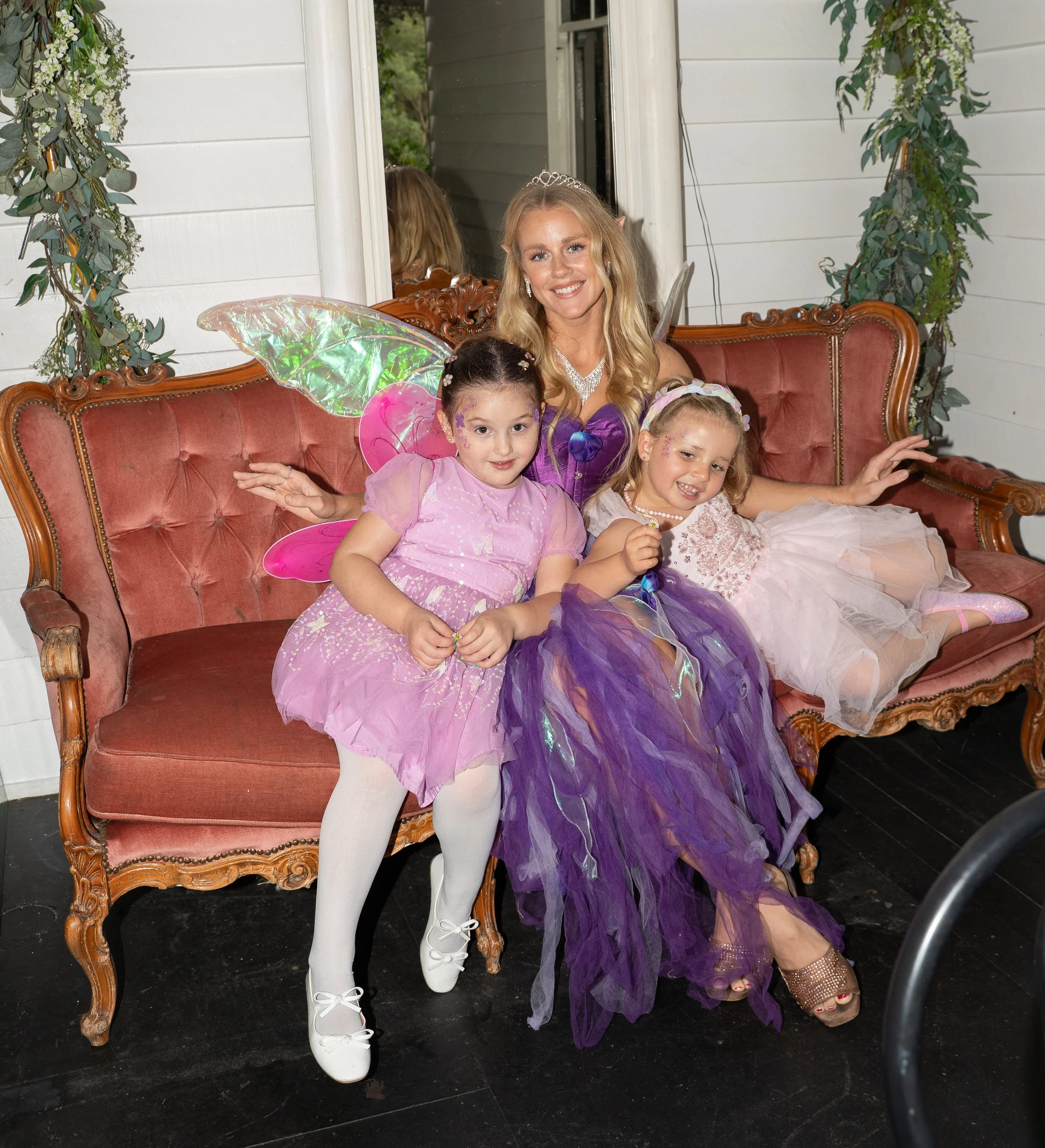 Three young girls dressed as fairies and a woman dressed as a princess or fairy sitting on a vintage red velvet sofa, with green foliage decorations around them, in an indoor setting.