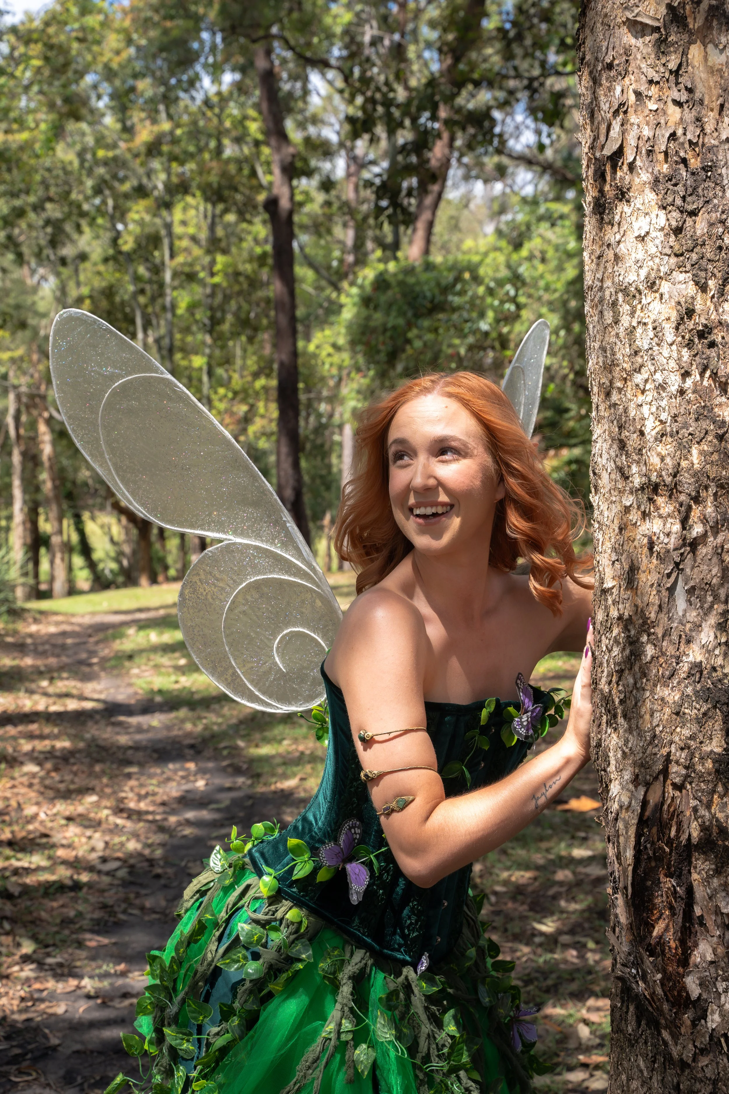 A young woman dressed as a fairy with butterfly wings and a green leafy dress is peeking out from behind a tree in a wooded outdoor setting, smiling and looking to the side.