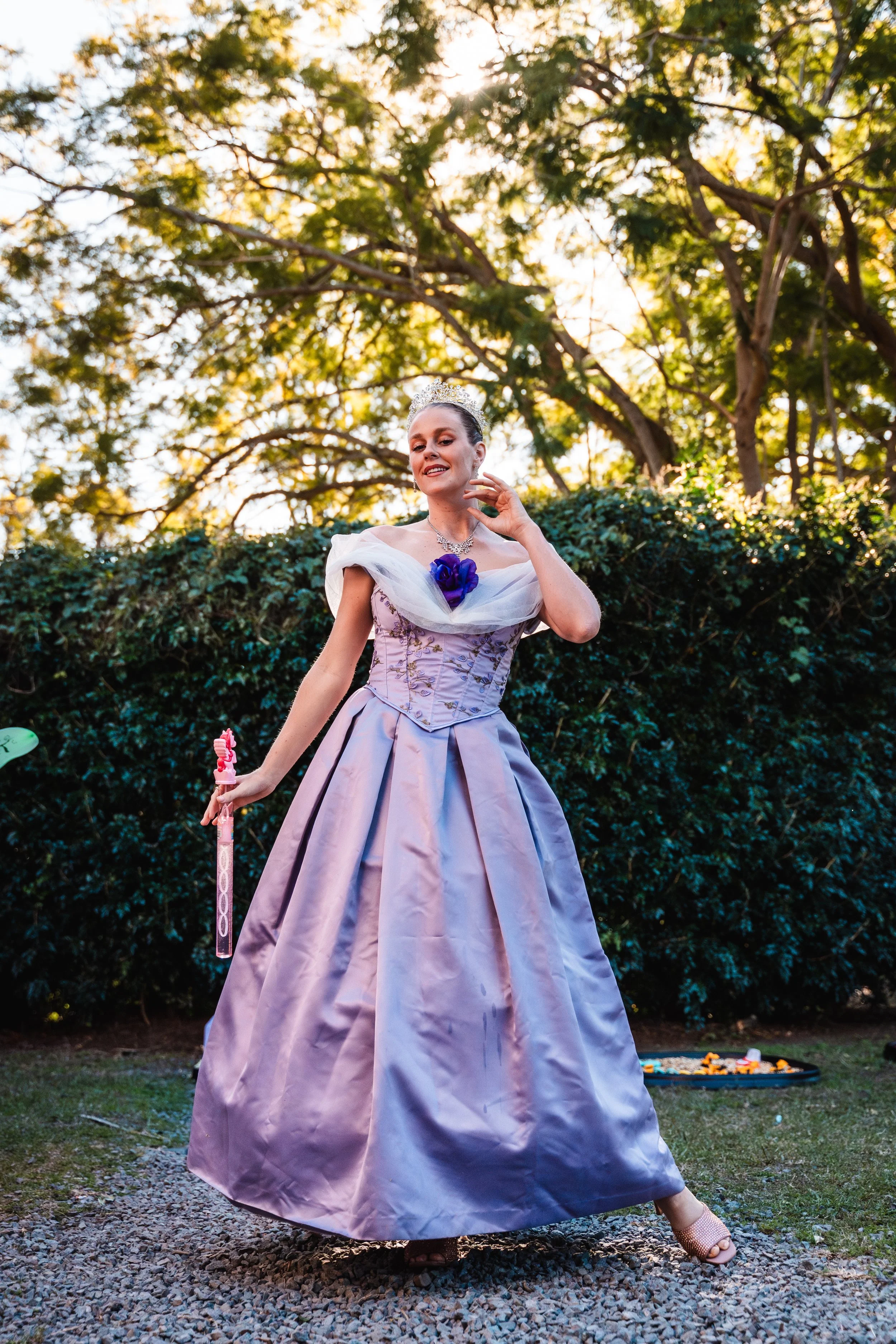 A woman in a lavender ball gown with puffed sleeves, wearing a tiara and necklace, holding a pink wand, standing outdoors on gravel with greenery and trees in the background, sun shining through the branches.