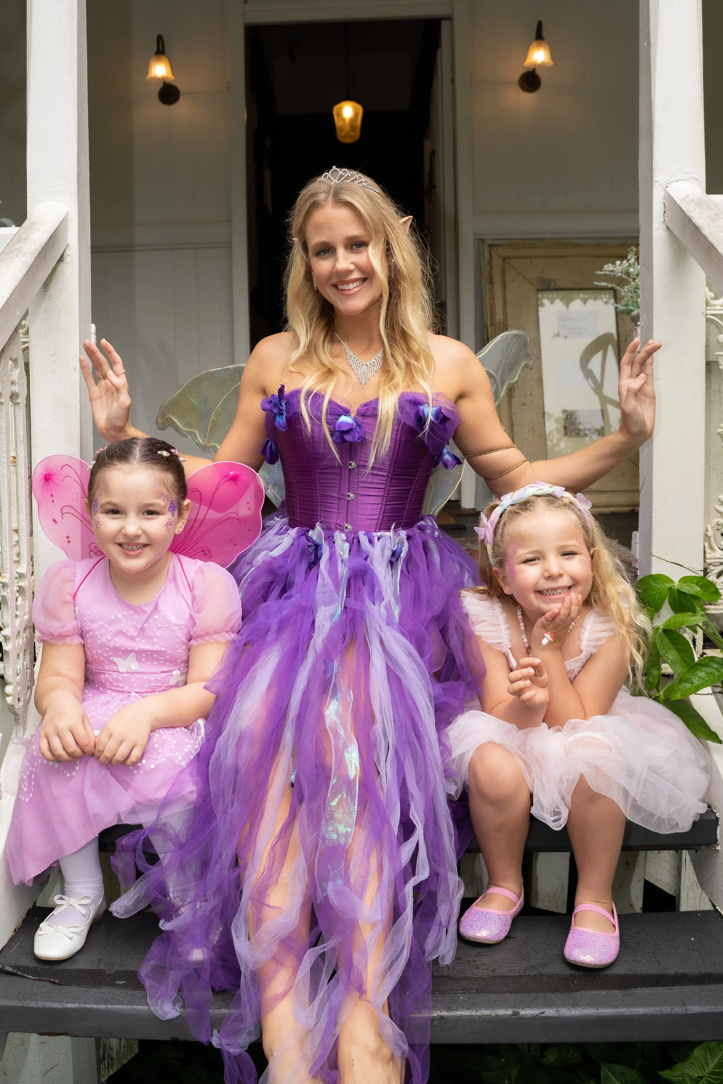 A woman dressed as a fairy with a purple dress, tiara, and butterfly wings, standing on a porch with two young girls in fairy costumes, one in pink and the other in light pink, smiling and posing for the camera.