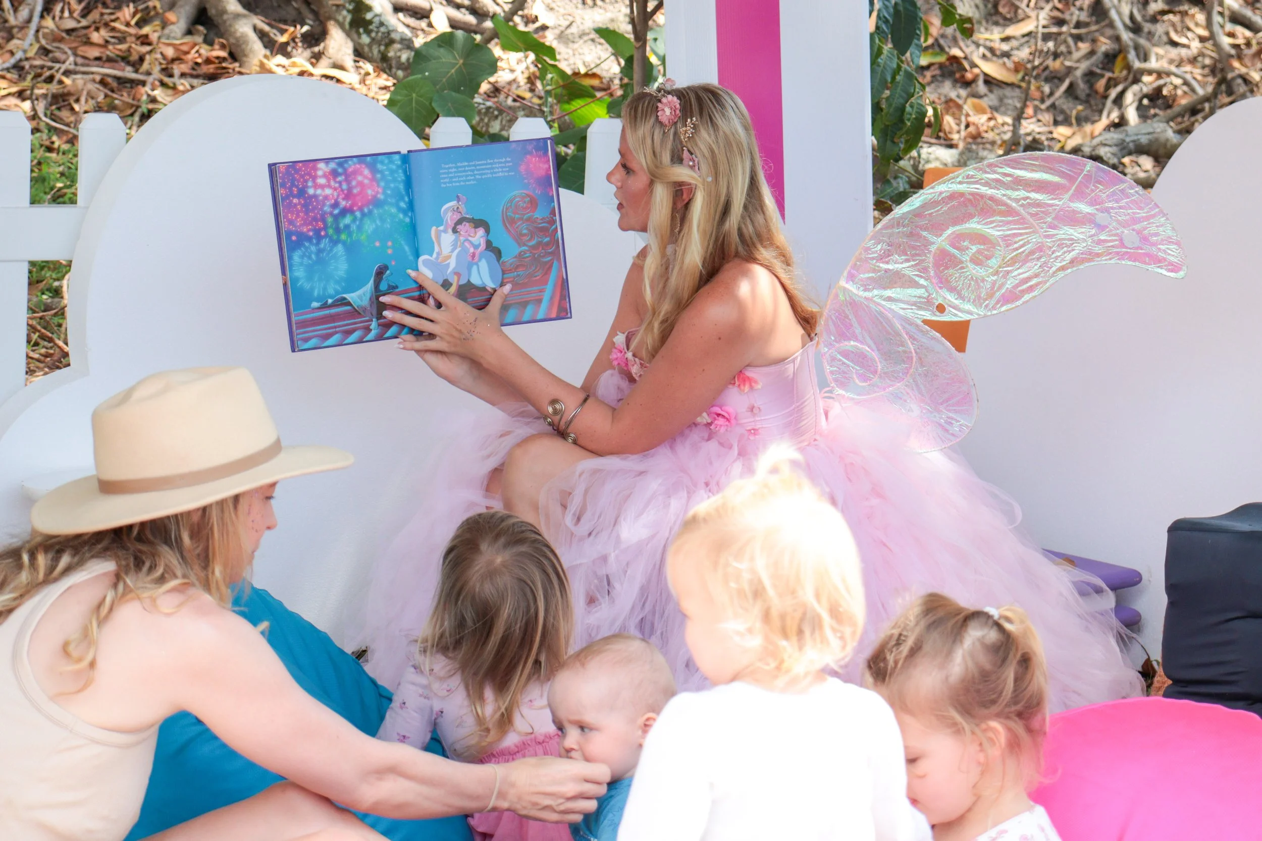 A woman dressed as a fairy princess reading a book to children at a children's party, wearing pink fairy wings and a pink fluffy dress, with children sitting around her.