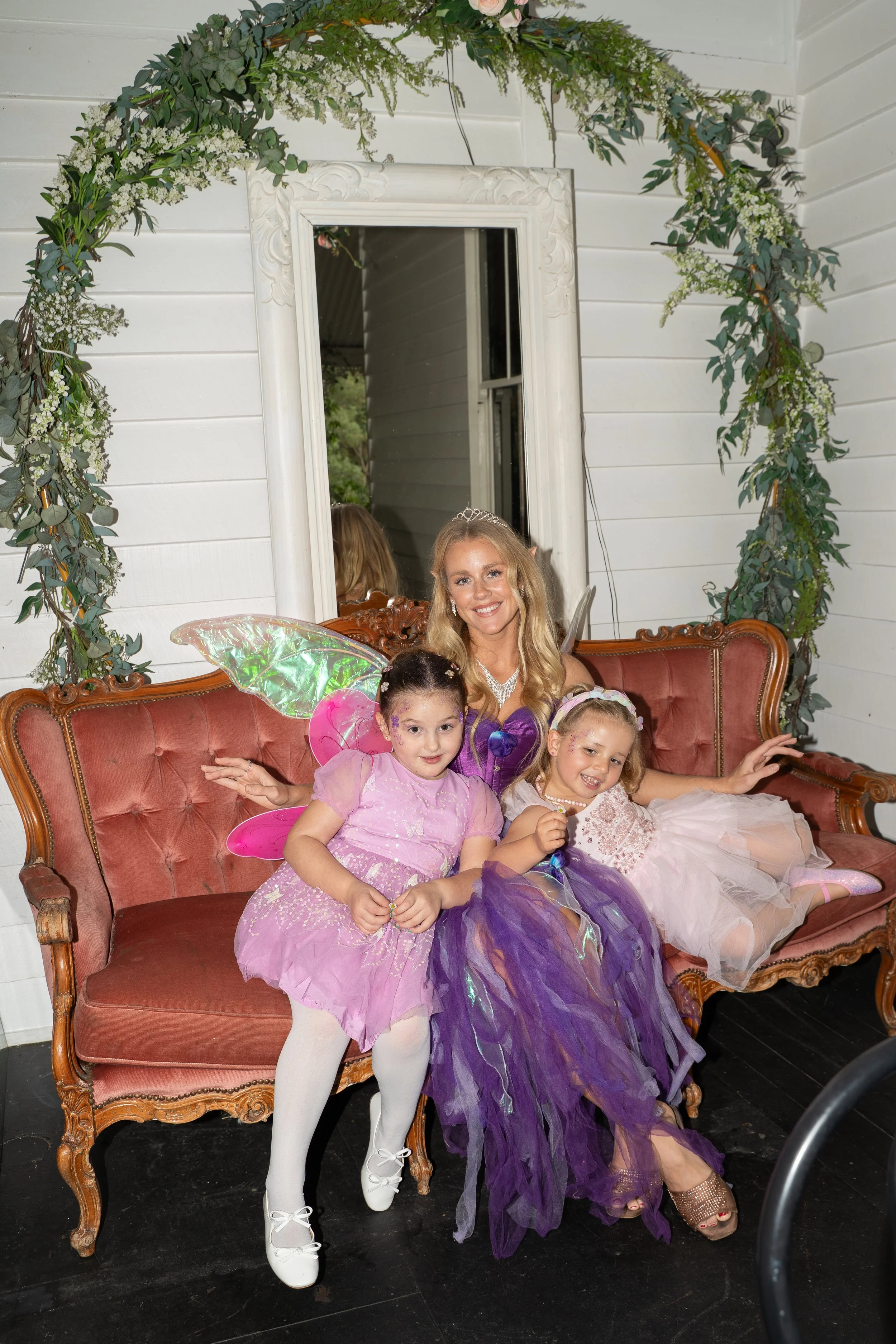 A woman dressed as a fairy princess, sitting on a vintage red sofa with three young girls dressed as fairies around her. The setting has a decorative mirror and floral greenery on the wall.