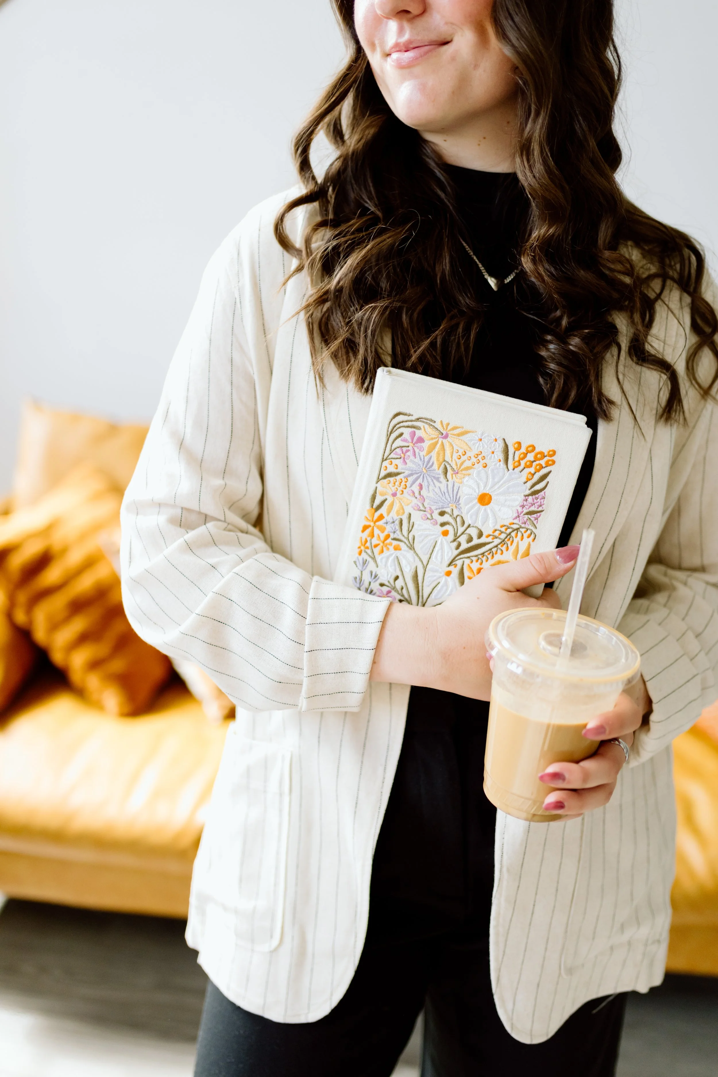 Woman with long, curly brown hair holding a floral embroidered notebook and iced coffee, wearing a cream-colored blazer with thin stripes, black top, and black pants, in a cafe or bakery setting with a yellow quilted sofa in the background.