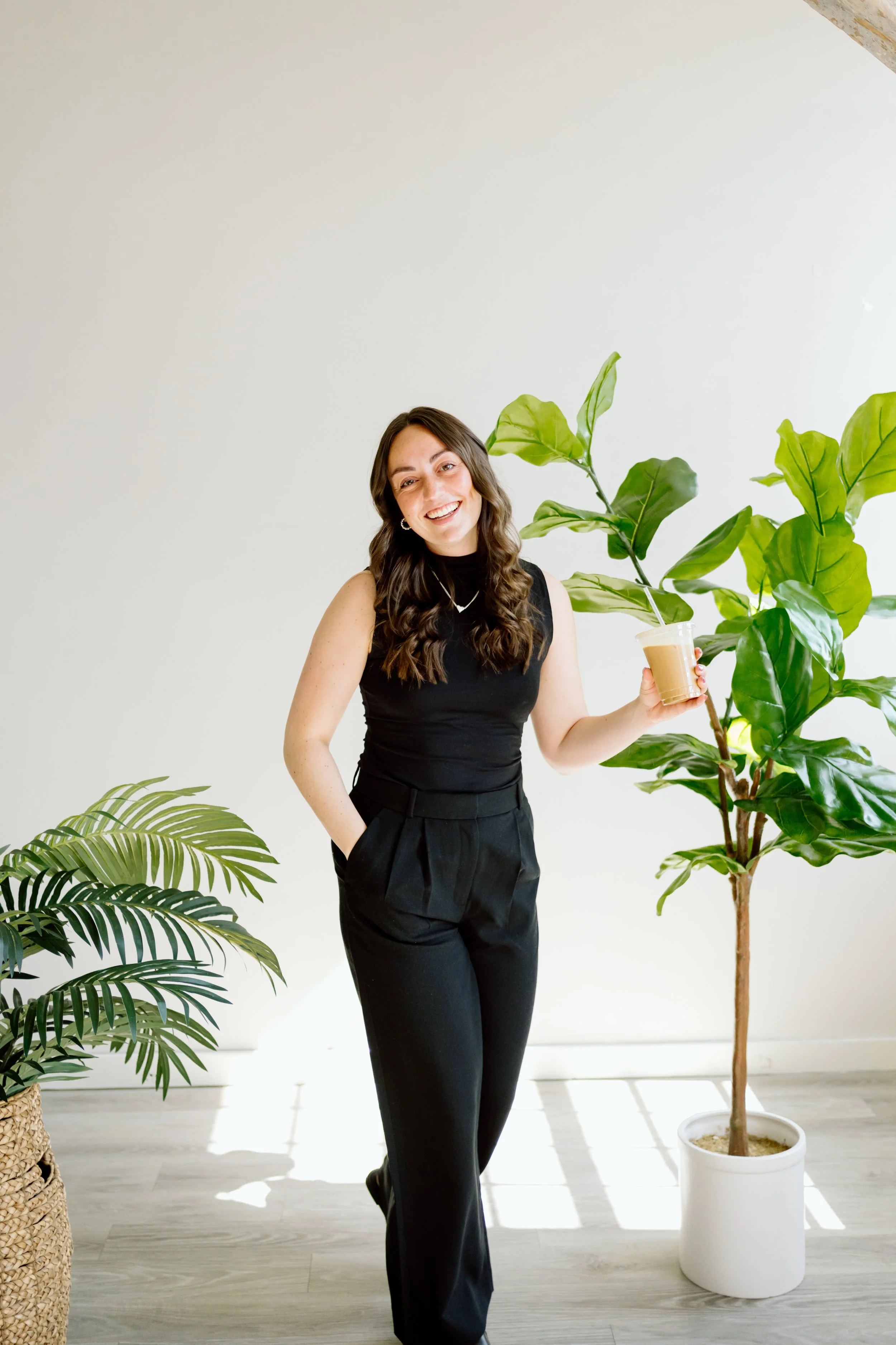 A woman with long brown hair wearing a sleeveless black turtleneck and black pants, smiling, holding a coffee drink in front of large green plants indoors.