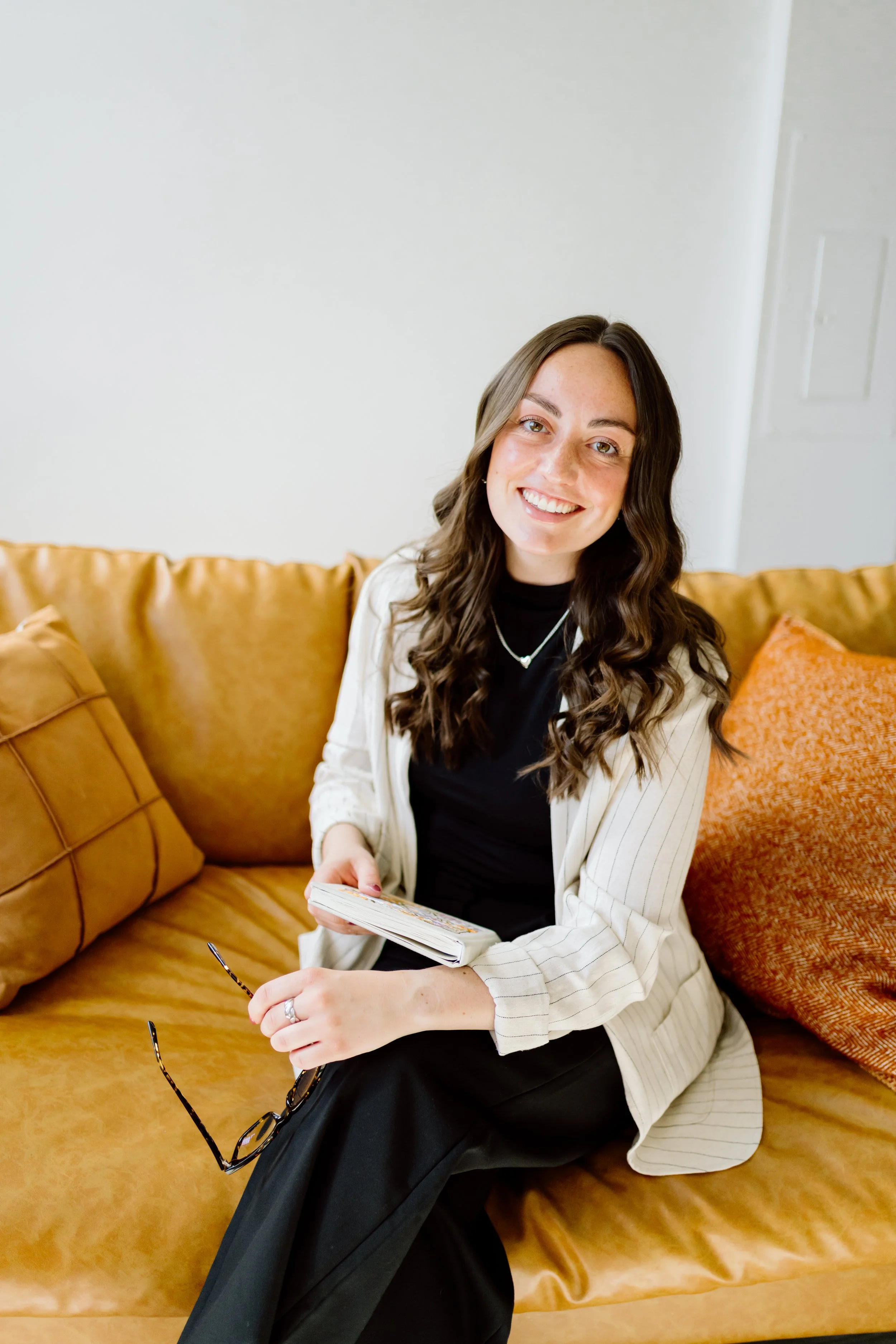 A woman with long wavy brown hair, smiling and holding a notebook, seated on a tan couch with matching cushions, in a room with white walls and a white door.