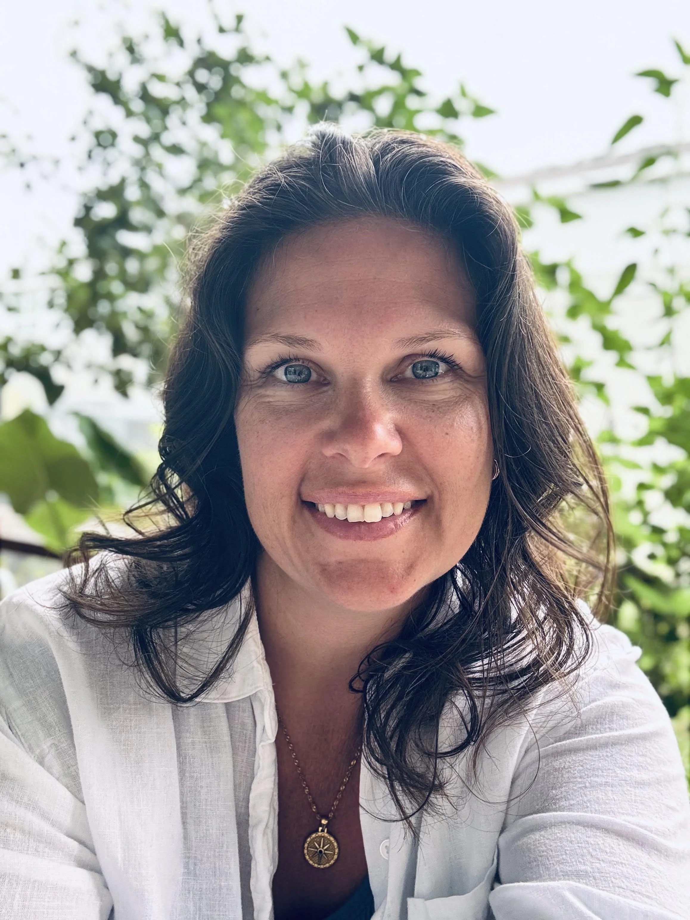 A smiling woman with dark, wavy hair and blue eyes, wearing a white shirt and a gold necklace with a compass pendant, outdoors with green leaves and sunlight in the background.