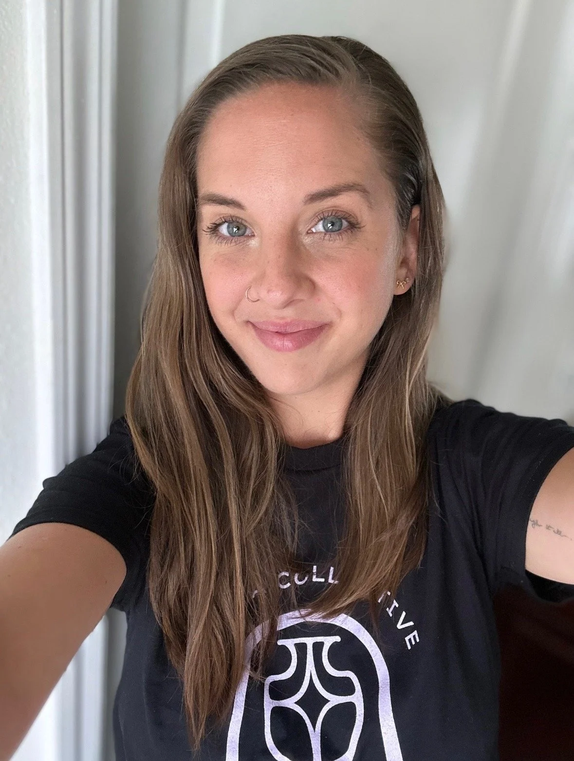 A young woman with long brown hair, blue eyes, wearing a black t-shirt with a logo, taking a selfie indoors near a white door.