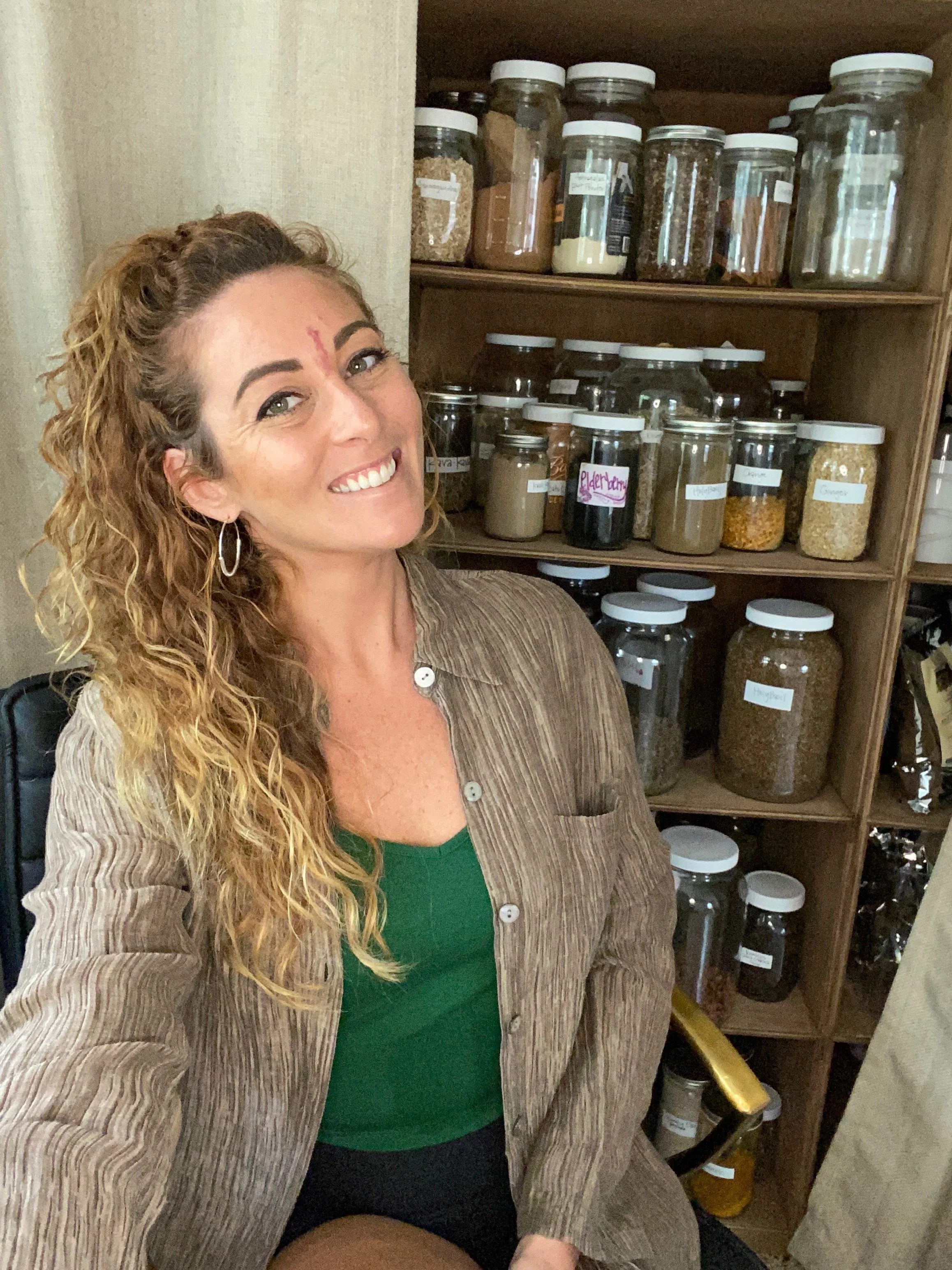 A smiling woman with curly blonde hair wearing hoop earrings, a green top, and a brown striped jacket, seated next to a bookshelf with various jars and containers filled with spices and herbs.