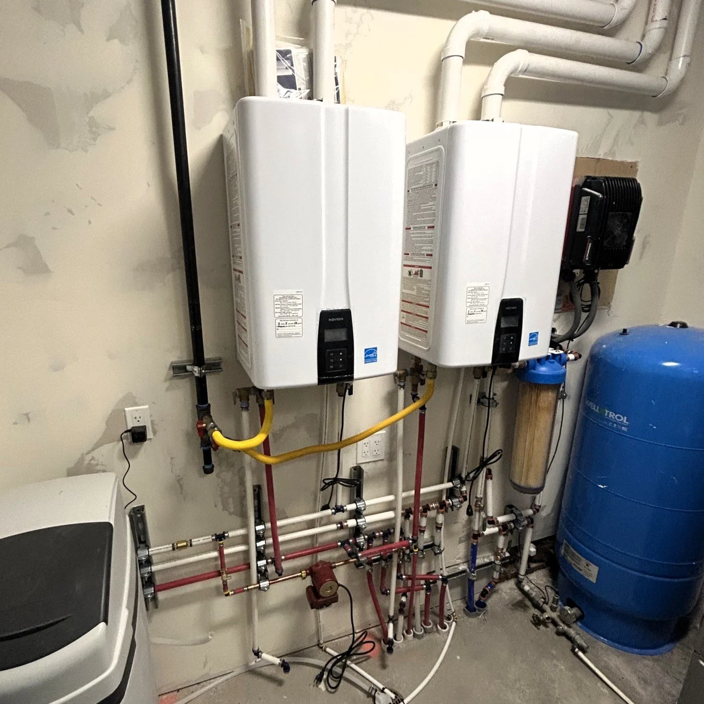 A utility room with two white tankless water heaters mounted on the wall, connected to various pipes, with a blue water pressure tank on the right and a washing machine partially visible on the left.