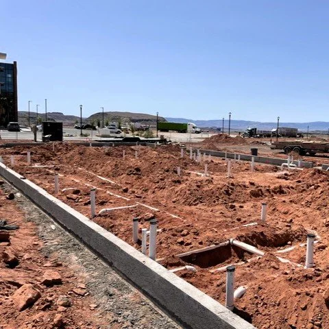 Construction site with exposed dirt, white pipes protruding from the ground, and a paved border along the site, with vehicles and buildings in the background under a clear blue sky.
