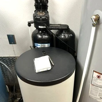 A large white water softener tank with a black lid, connected to two black tanks and a gray water heater in a utility room.