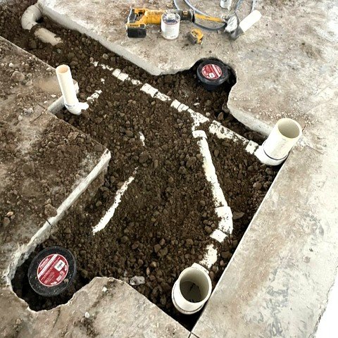 Underground plumbing pipes with white PVC pipes and fittings installed in an excavated trench, surrounded by dirt and construction tools.