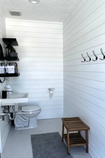 A minimalist bathroom with white shiplap walls, a small white sink, a wall-mounted toilet, black shelving holding plants and towels, a wooden stool, black hooks on the wall, and a gray rug.