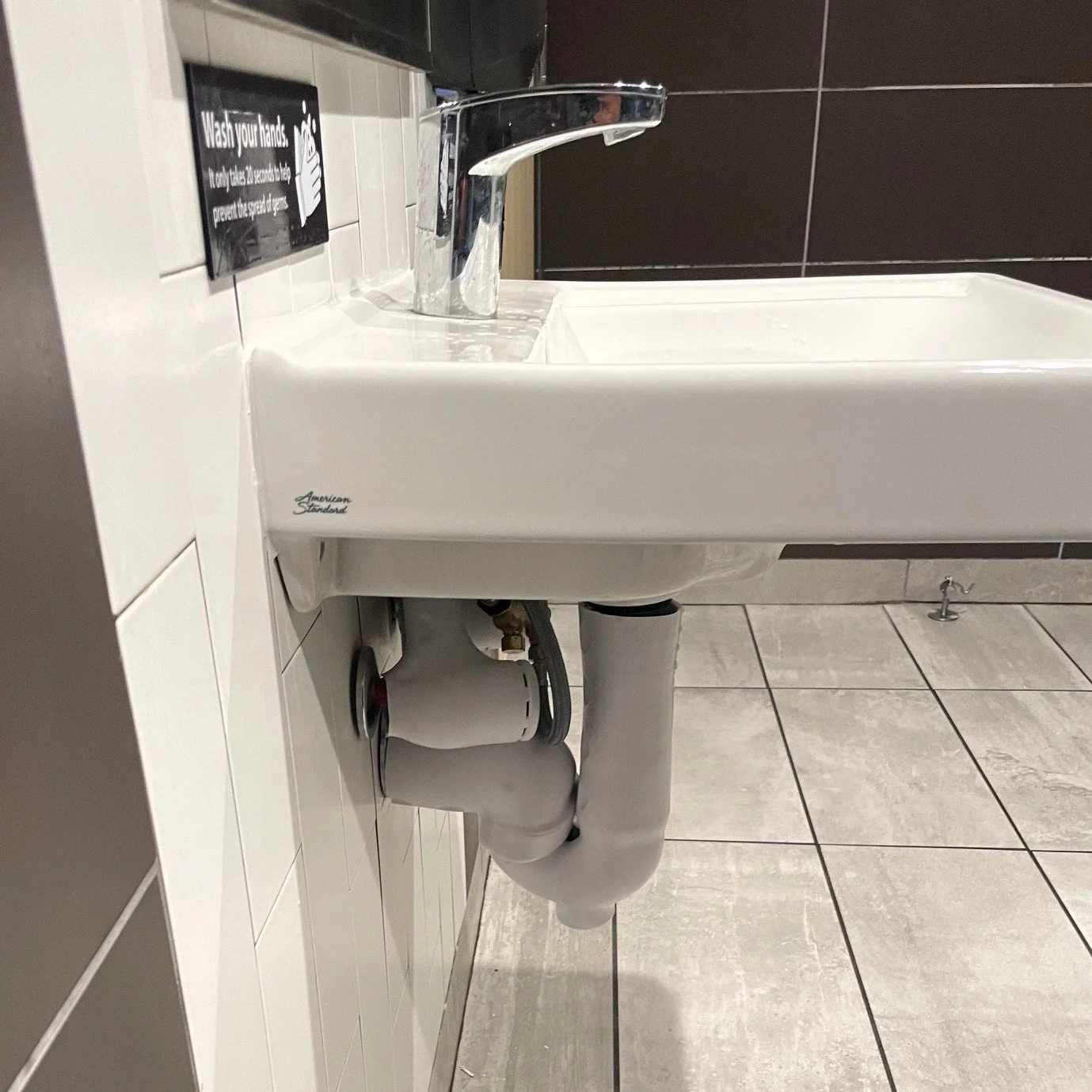 A white sink with a chrome faucet mounted on a white tiled wall with a black and white wash your hands sign nearby. The sink has visible plumbing underneath and beige tiled floor.