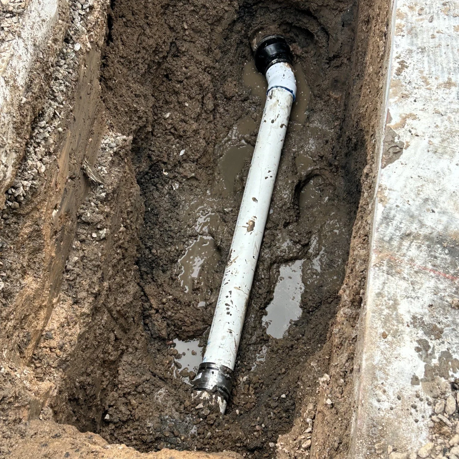 A hole dug in the ground shows white PVC piping connected at the bottom, with muddy water surrounding it.