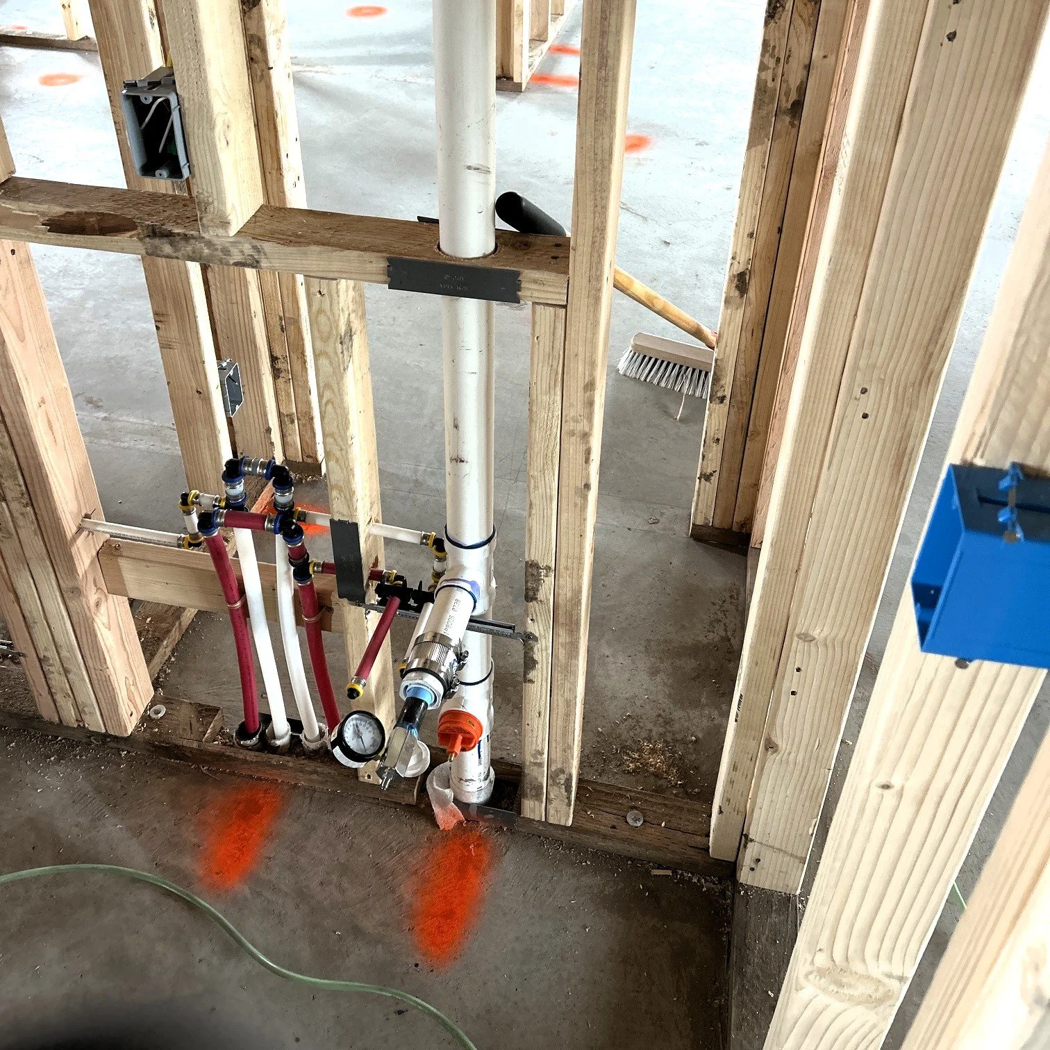 Close-up view of plumbing and piping system installed in wall framing of a building under construction. Includes white and red pipes, a pressure gauge, and an orange valve, with wooden studs and concrete floor visible.