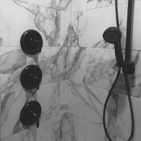 A marble tiled shower wall with three black round shower controls and a black handheld shower head attached to a hose.