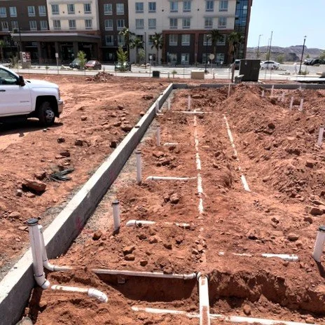 Construction site with exposed pipes and outlines for building foundation, with cars and apartments in the background.