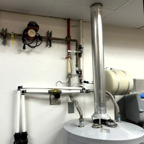 Piping and ductwork in a mechanical room, including a vertical metal vent pipe, valves, gauges, and control components.