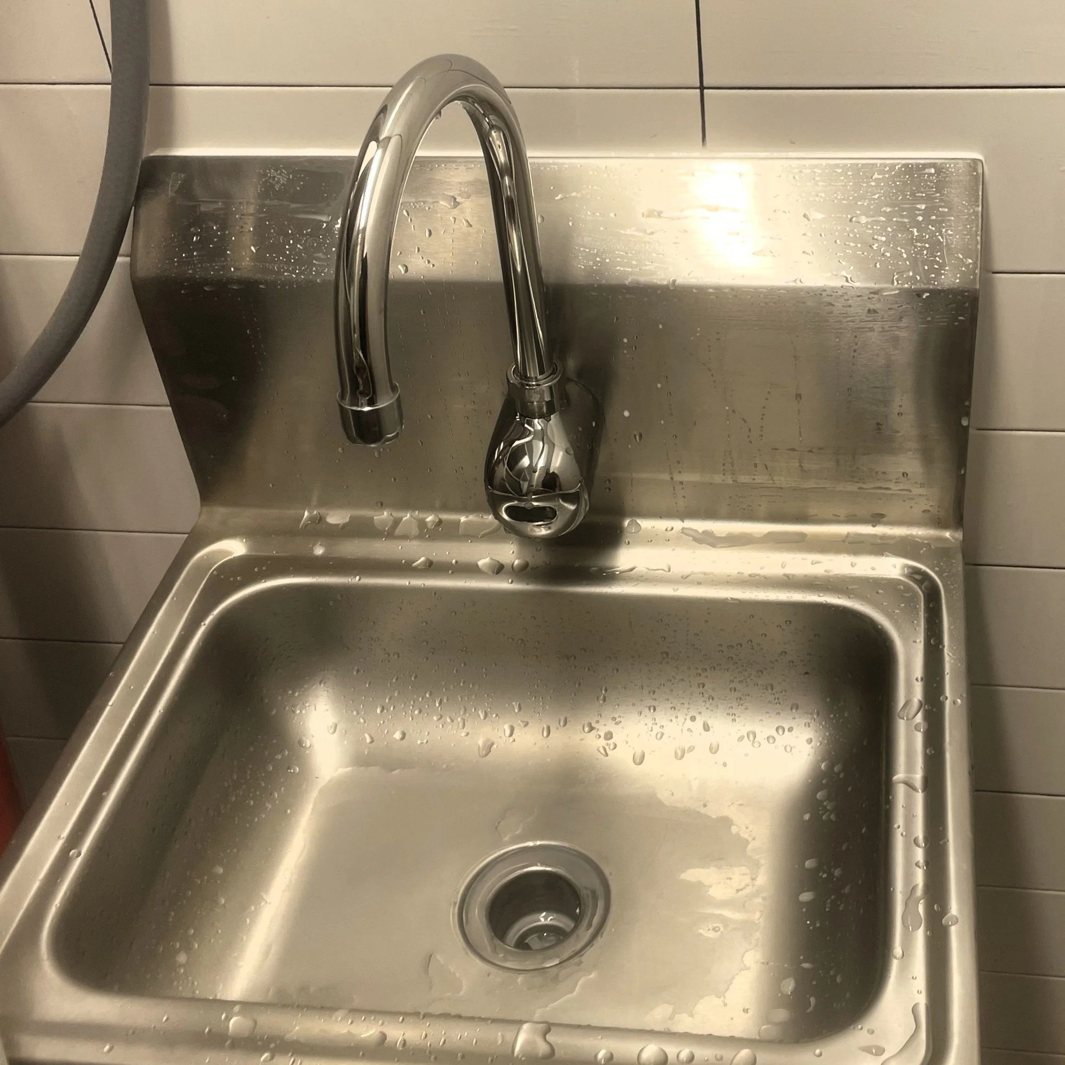 A stainless steel kitchen sink with a faucet, water droplets on the surface, and a tiled wall background.