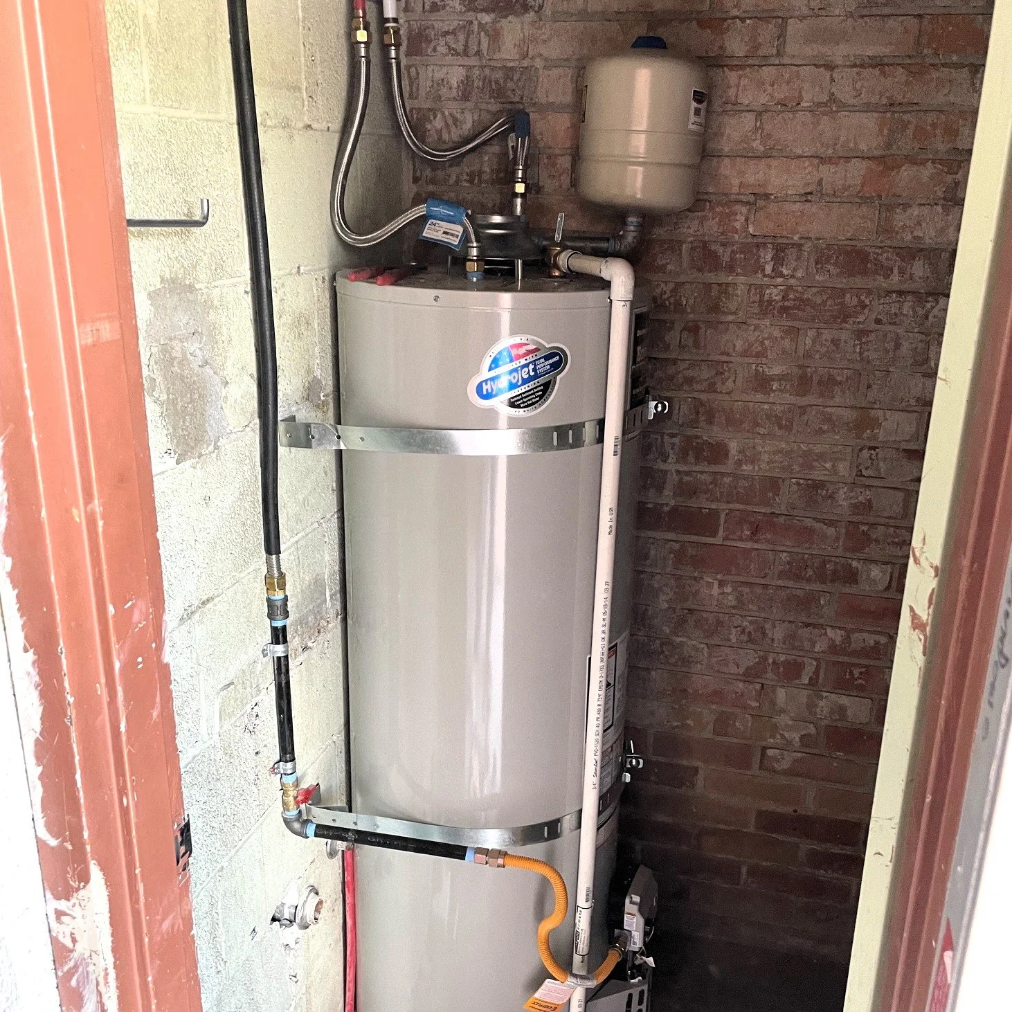 A white water heater installed in a small utility closet with brick wall background, connected to various pipes and hoses.