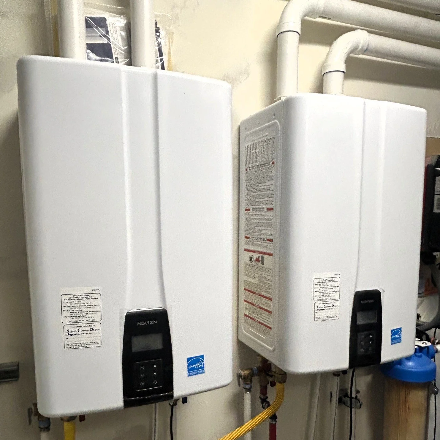 Two white tankless water heaters mounted on a beige wall with visible pipes and labels.