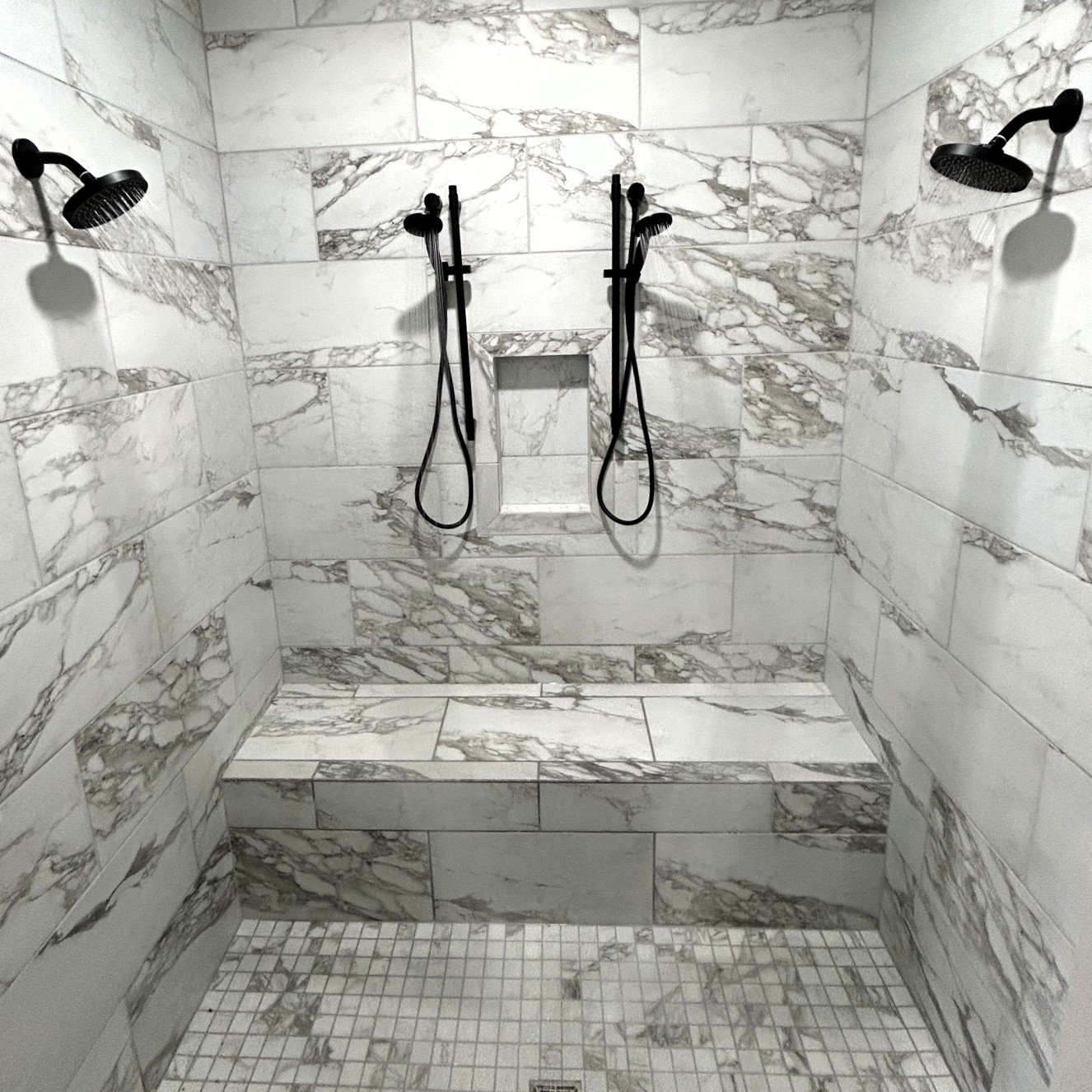 A walk-in shower with marble tile walls, two black showerheads on each side, a built-in niche for toiletries, and small square tile flooring.