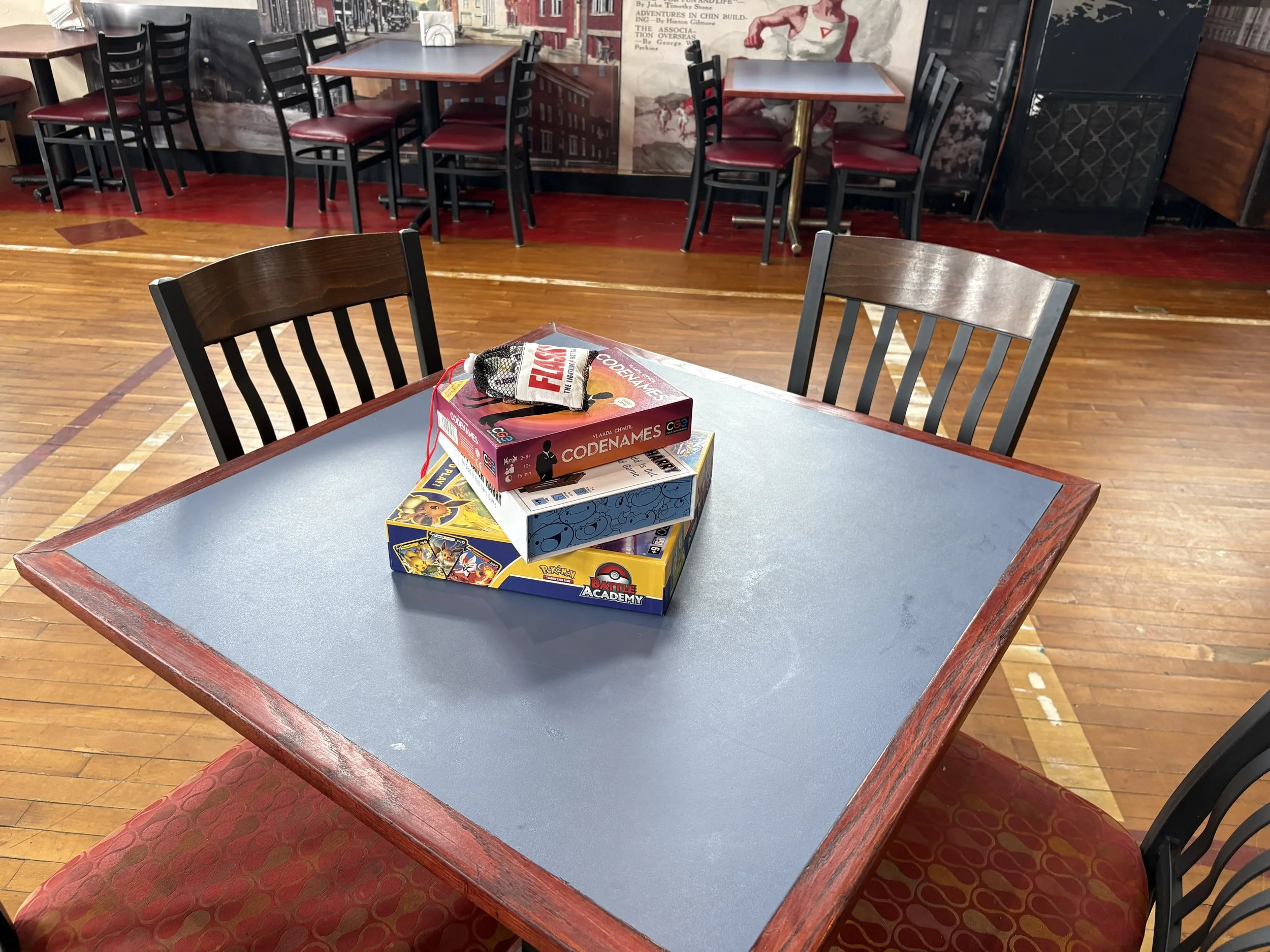A tabletop with boxed board games and a small cloth bag. In the background, there are multiple tables and chairs in a restaurant or cafe setting, with a mural or poster on the wall depicting historical images and text.