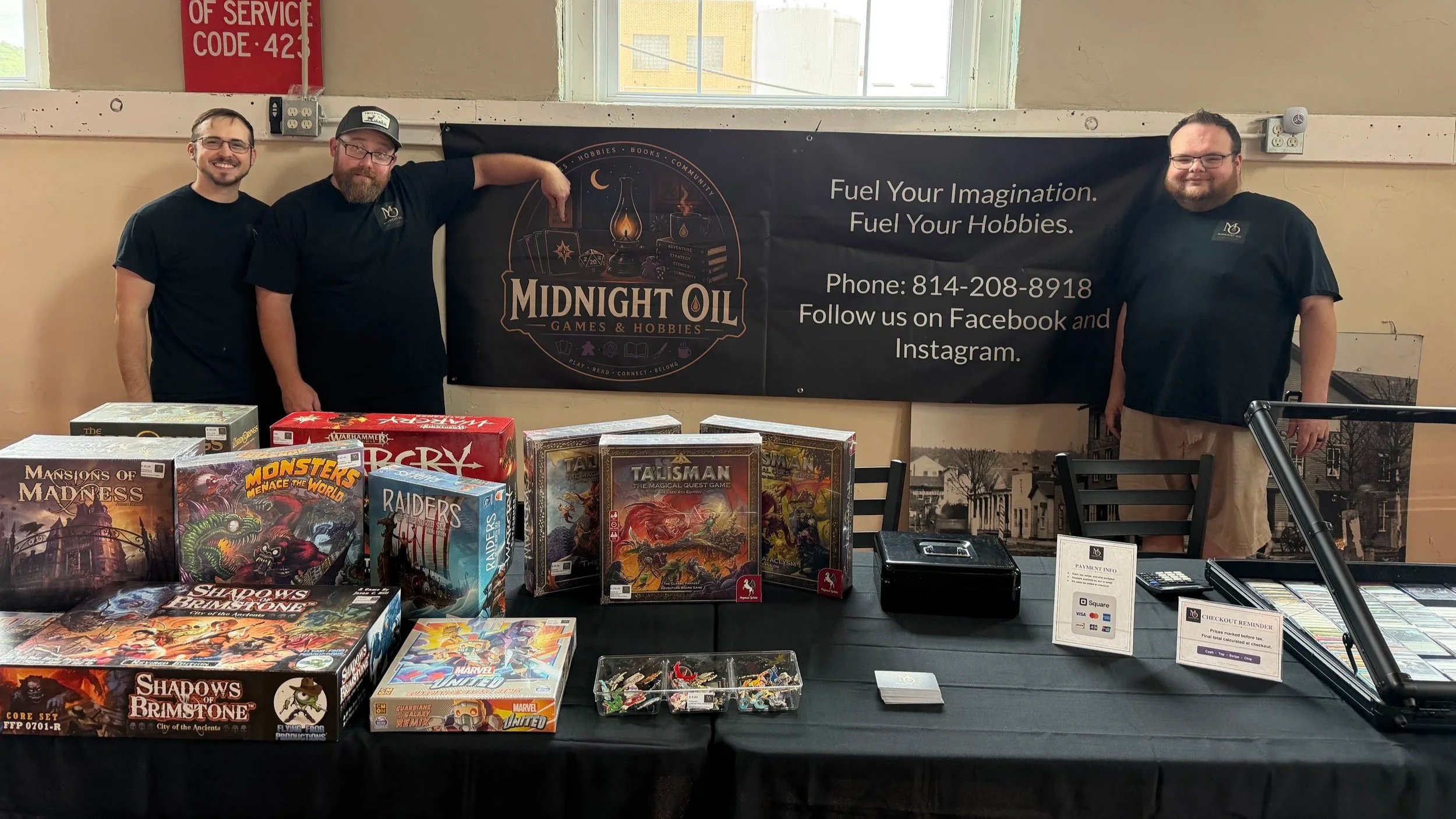 Three men standing behind a table with board games at a gaming event. One man points to a banner with the Midnigh Oil Games & Hobbies logo, and the table displays various game boxes like Shadows of Brimstone, Monster Menace, and Raiders of the Lost Sea.