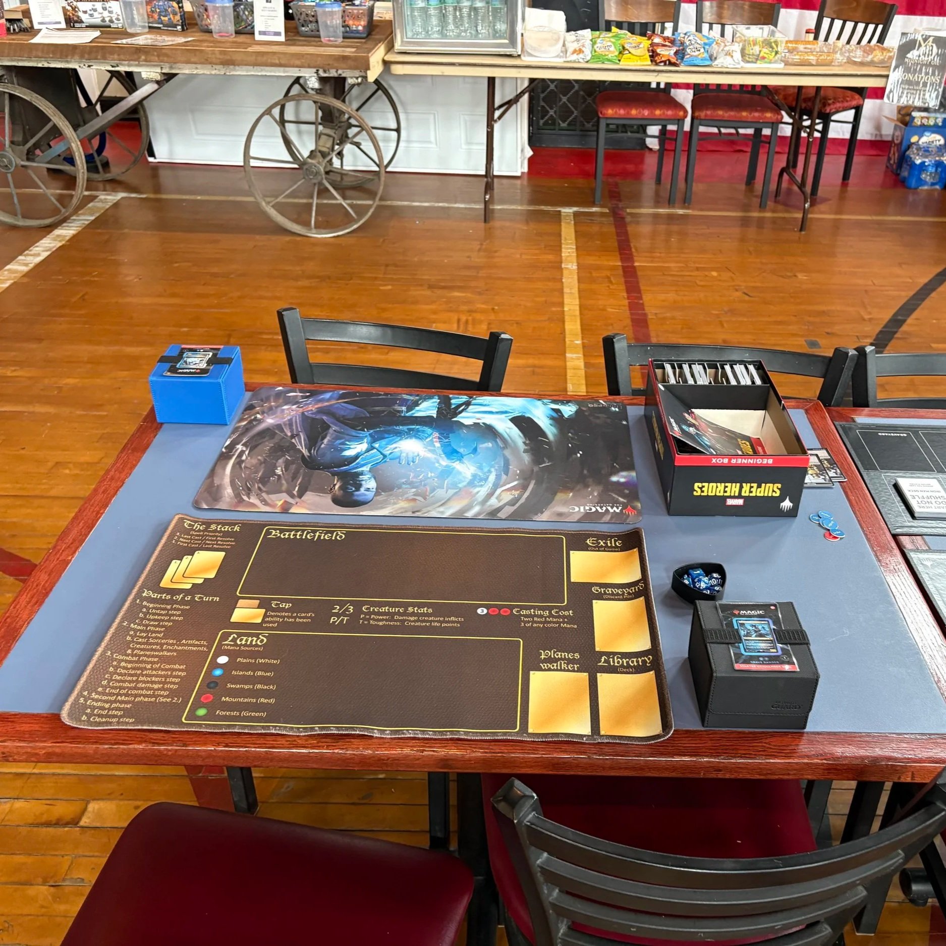 A tabletop strategy game setup with a game rulebook, a deck of cards, dice, and game pieces. The background shows chairs, a counter with snacks, and a fridge in a recreational or game room setting.