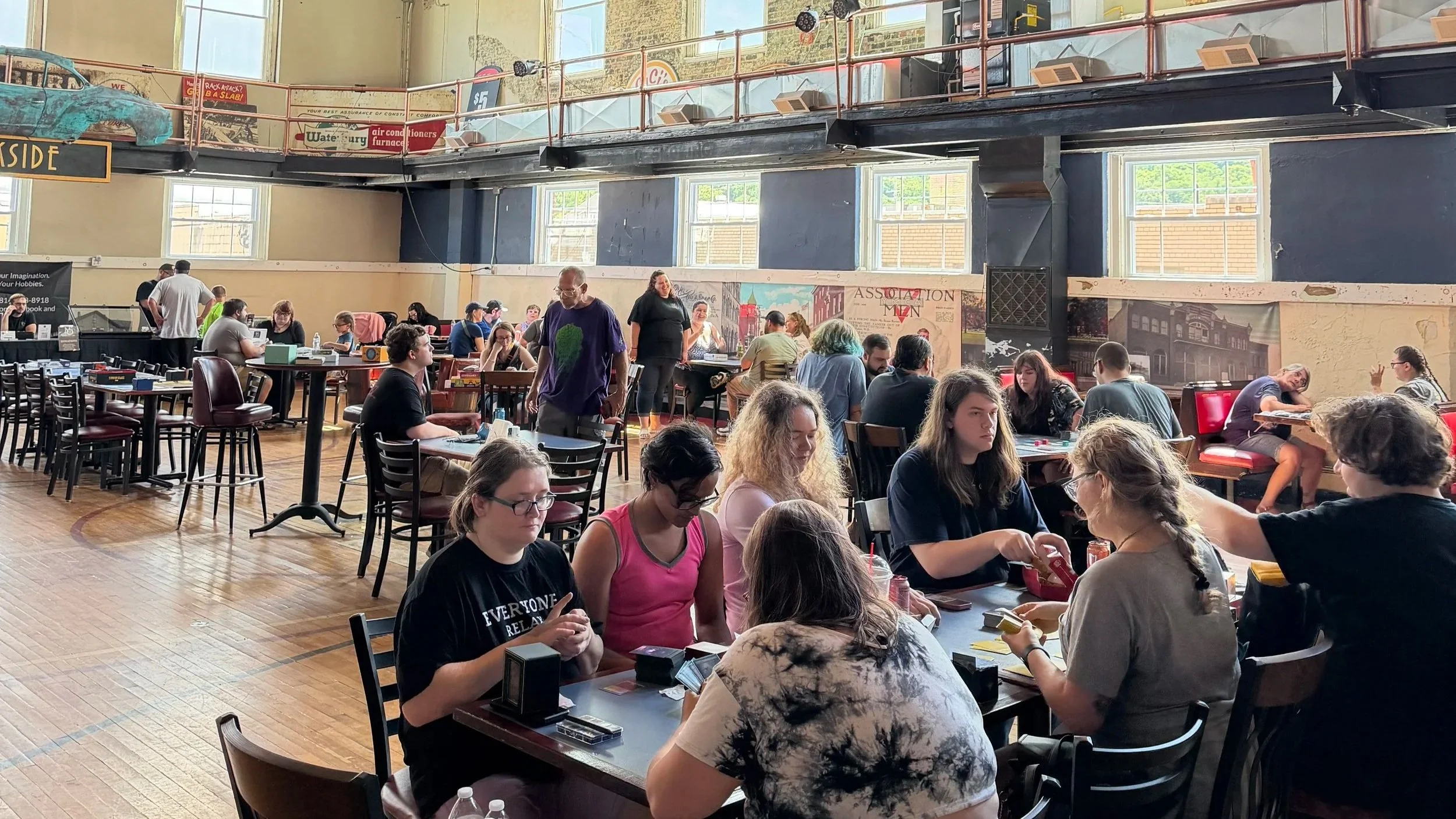 People playing a card game at tables in a large indoor space with high windows, a balcony, and vintage decor, resembling a game store or event venue.