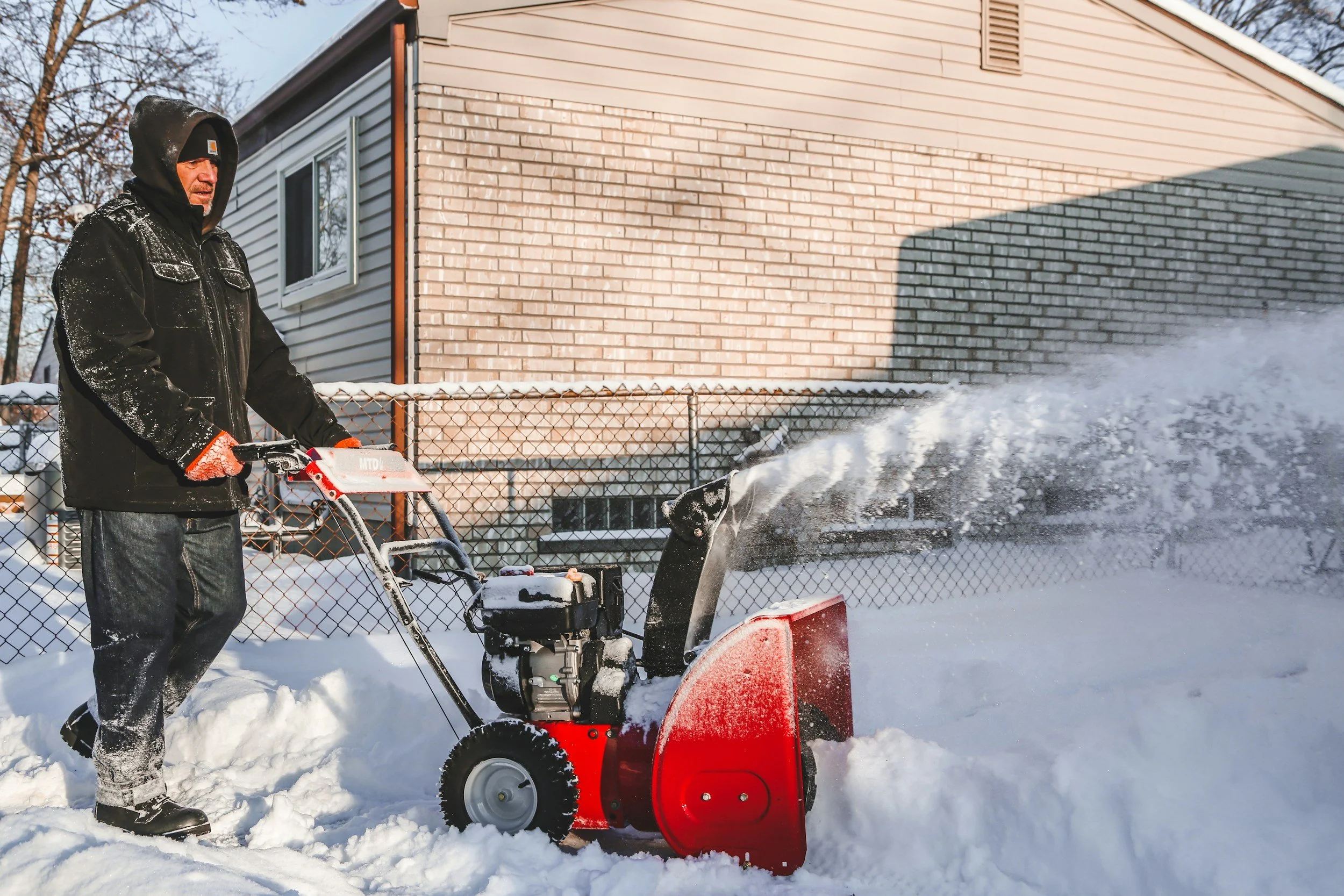 A man wearing a black hooded jacket and orange gloves is operating a red snowblower on a snow-covered driveway, with a house and a fence in the background.