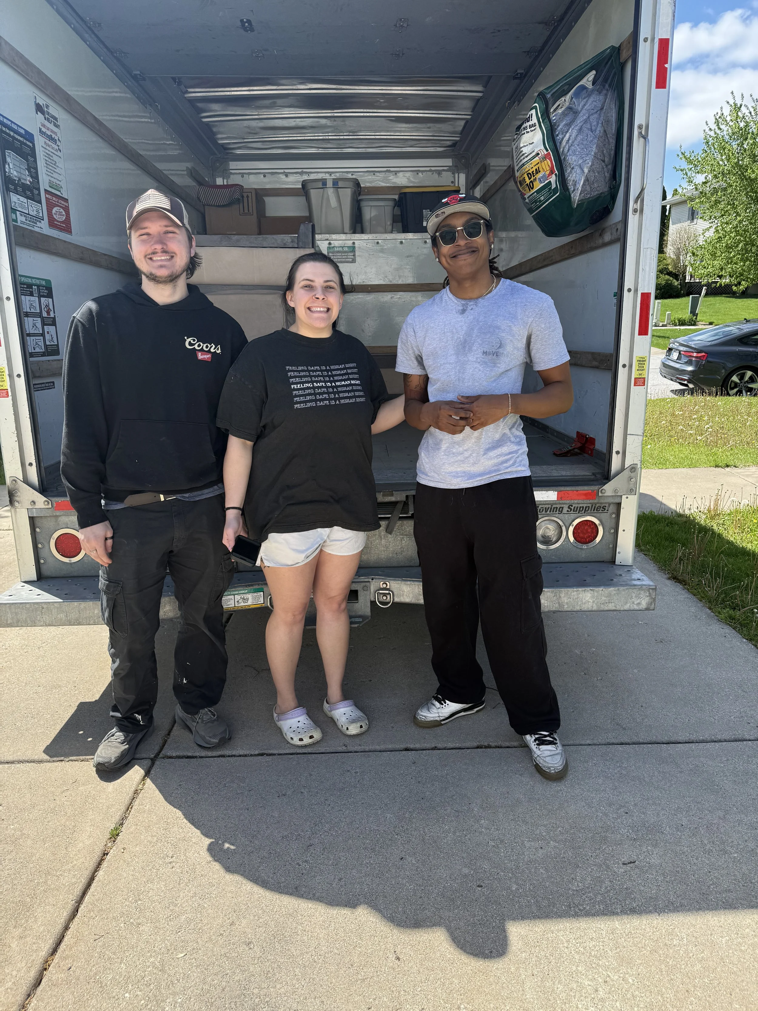 Three smiling people standing in front of an empty moving truck, with a green green bag hanging on the right side of the truck. The background shows a suburban neighborhood with cars parked on the street and houses with trees.