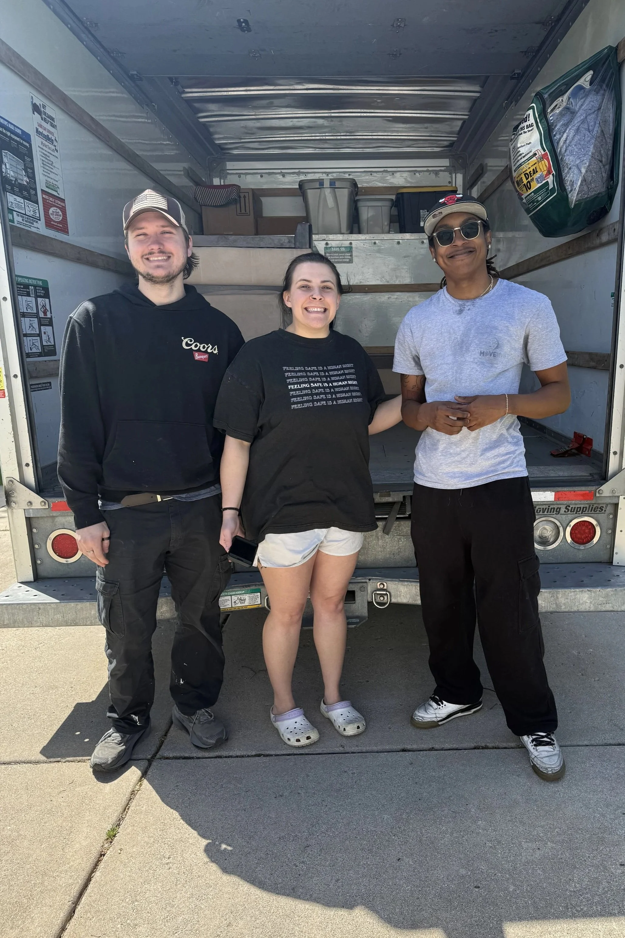 Three smiling people stand in front of an open moving truck, posing for a photo on a sunny day.