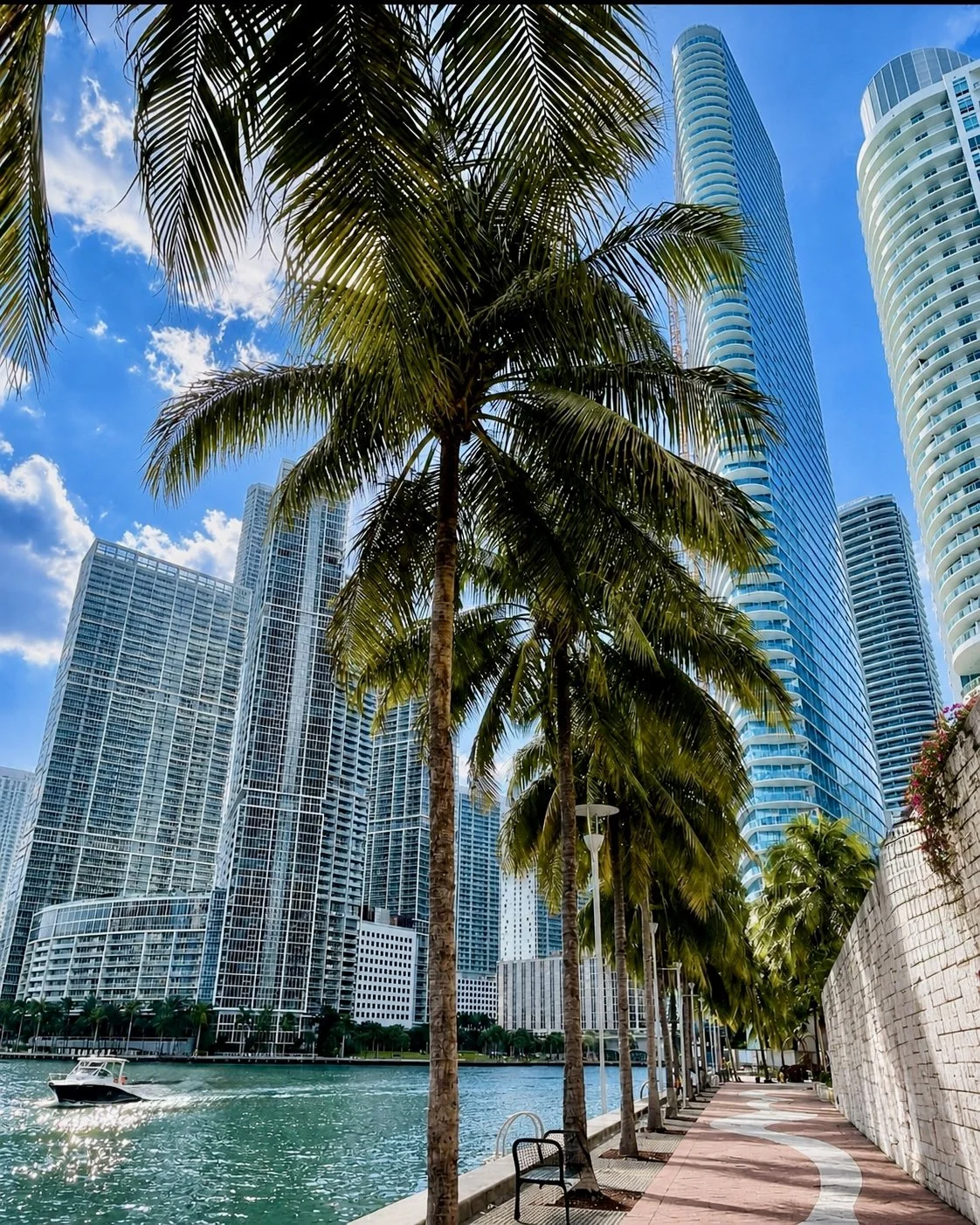 A Brickell, Biscayne Bay walk, Miami with tall modern skyscrapers, a waterfront, and palm trees along a pathway, under a blue sky with clouds.