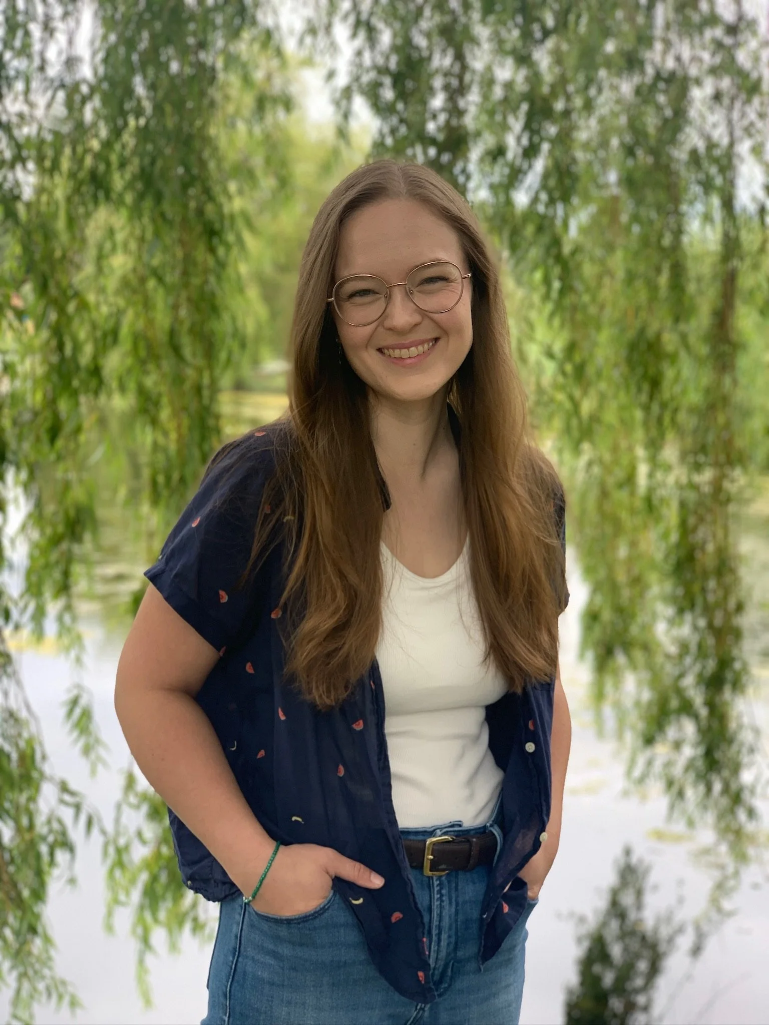 A young woman with long brown hair, glasses, and a friendly smile standing outdoors in a lush, green environment with trees and water in the background.