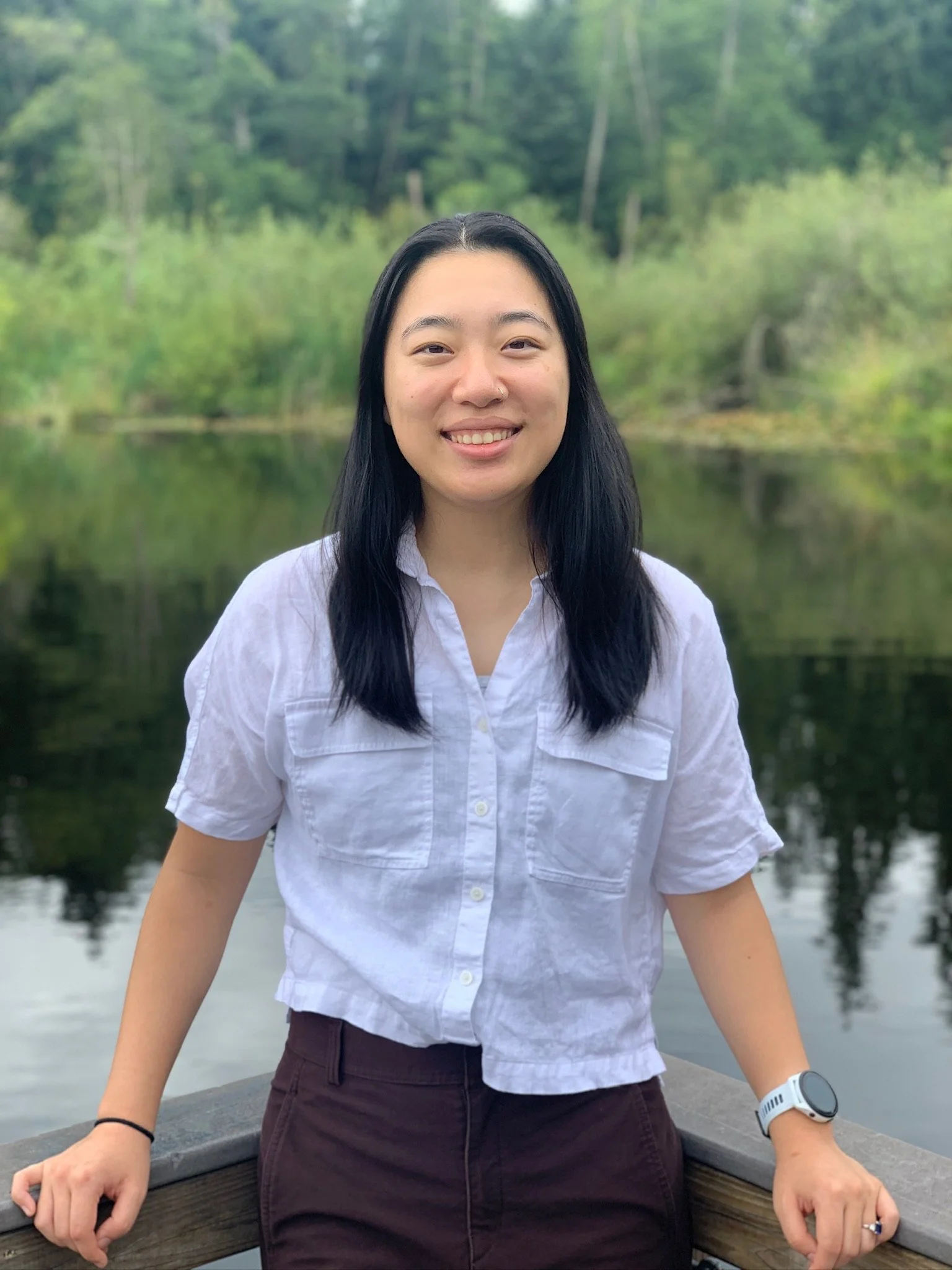A woman with long black hair smiling in front of a blurry green forest and a body of water, sitting on a boat or dock with her arms resting on a wooden railing.