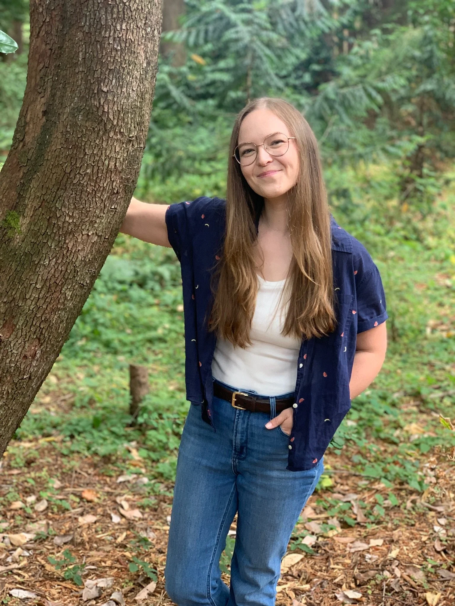 A woman with long brown hair and glasses standing in a wooded area, leaning against a tree with her left arm, smiling at the camera. She is wearing a white shirt, a dark blue shirt with small patterns, blue jeans, and a brown belt.