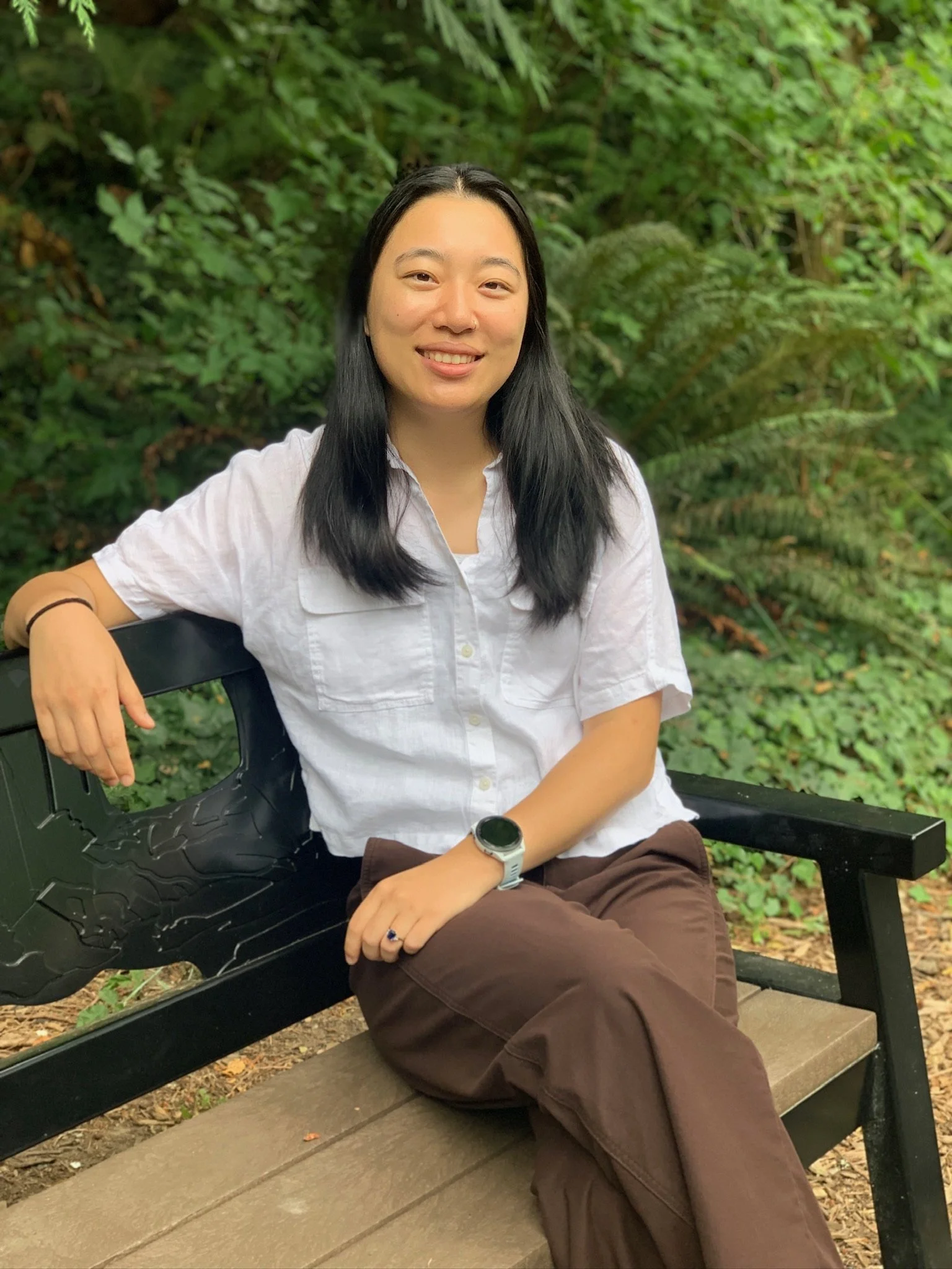 A young woman with long black hair sitting on a park bench in front of a green leafy background. She is smiling and dressed in a white short-sleeve button-up shirt and brown pants, wearing a wristwatch and a ring.