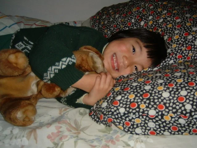 A young girl lying in bed, smiling, holding a plush dog toy, with colorful patterned pillows in the background.