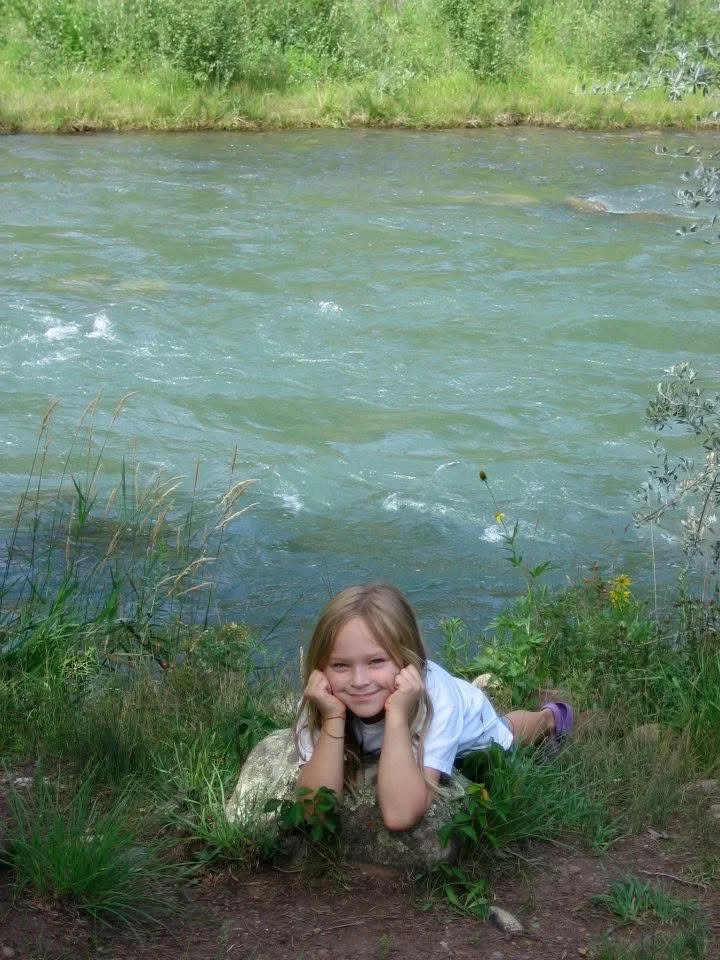 A young girl lying on her stomach on a rock near a riverbank, smiling and resting her chin on her hands, surrounded by green grass and plants.
