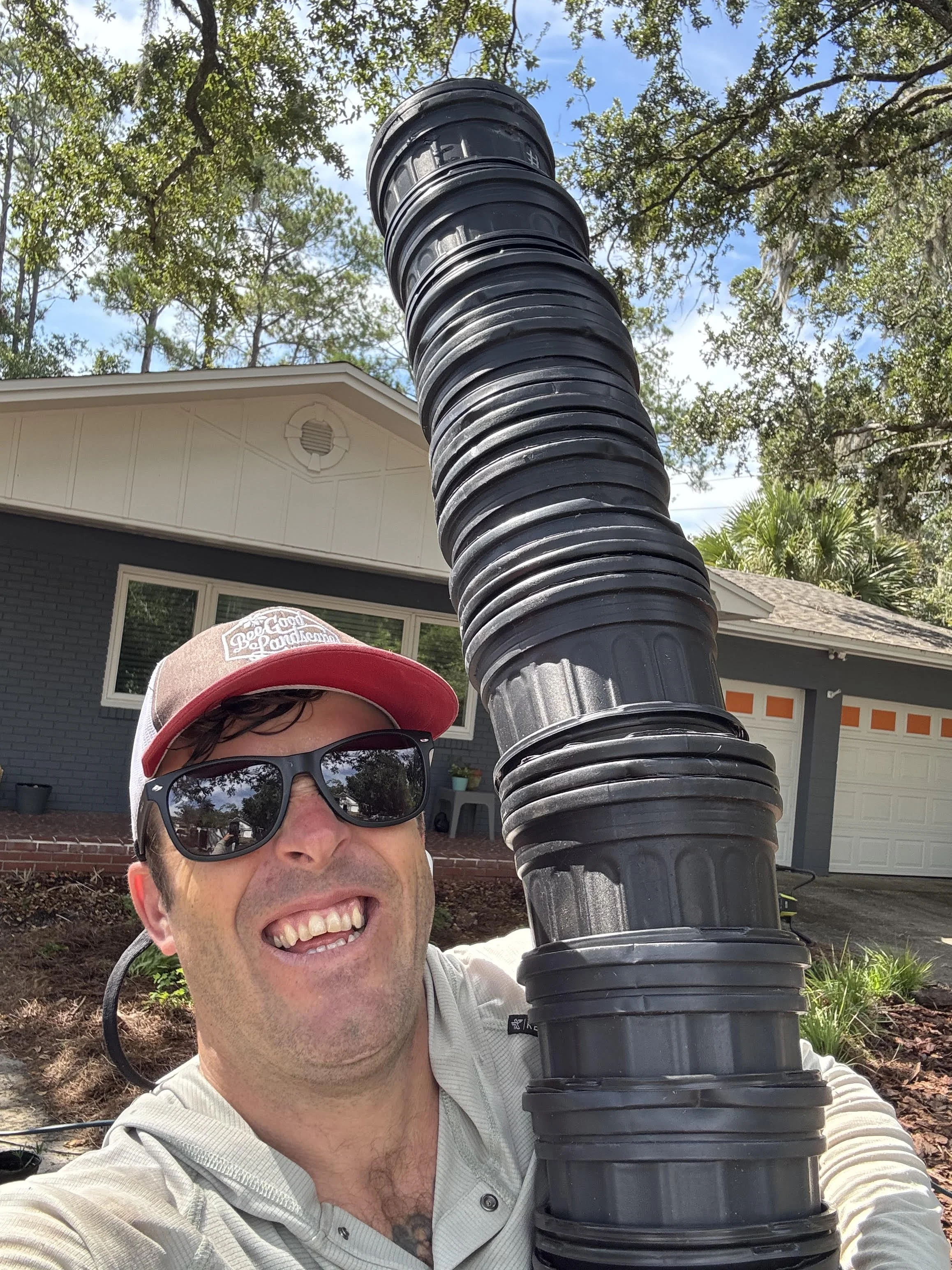 A man smiling, wearing sunglasses and a red cap, taking a selfie in front of a garage, with a large black plastic pipe or tube in the foreground, and green trees and a house in the background.