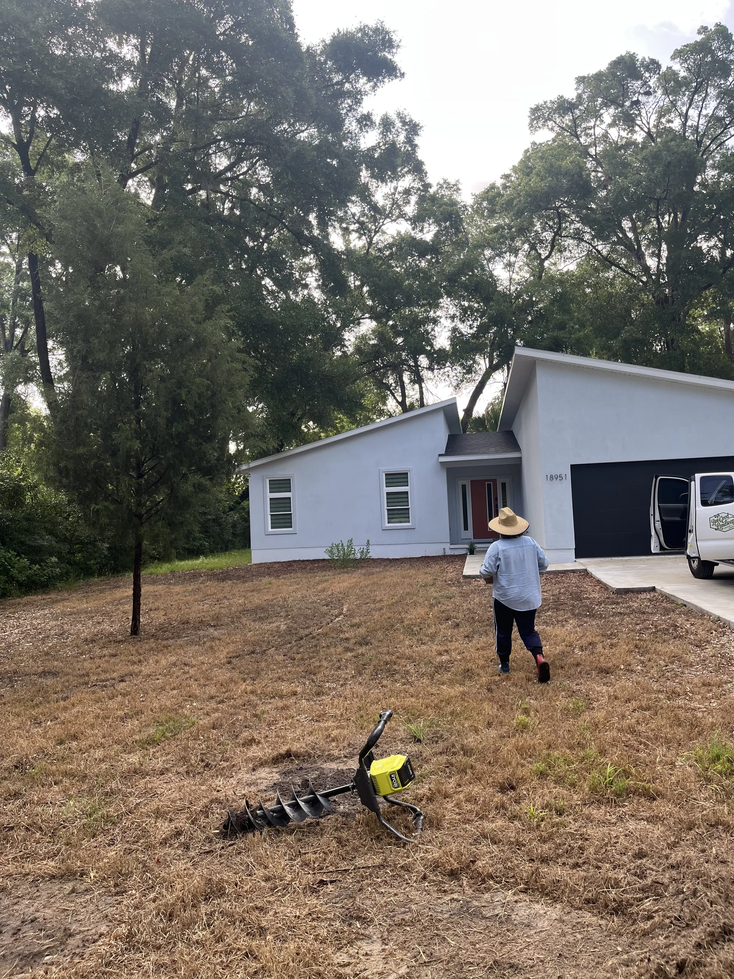A person wearing a wide-brimmed hat, white shirt, and black pants walking across a yard with dried grass, beside a black and yellow auger tool lying on the ground. A white house with a dark garage door and large trees with green leaves in the background.