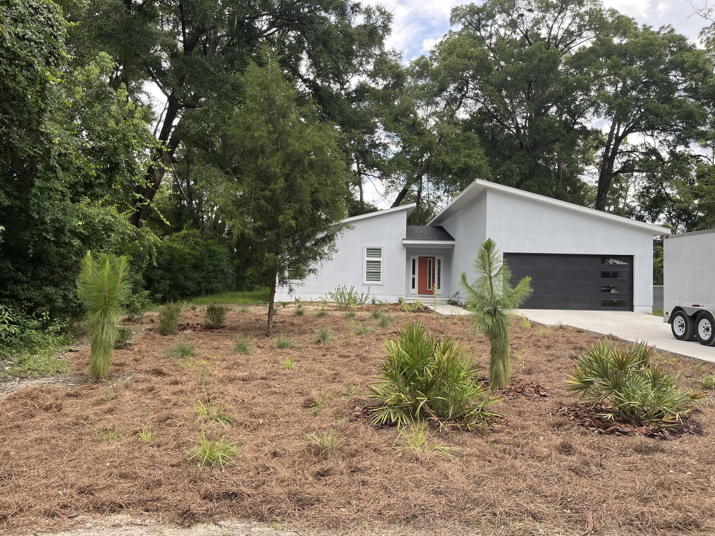 A modern white house with a black garage door and a small front porch, surrounded by trees and a lawn with sparse plants and patches of dry ground.